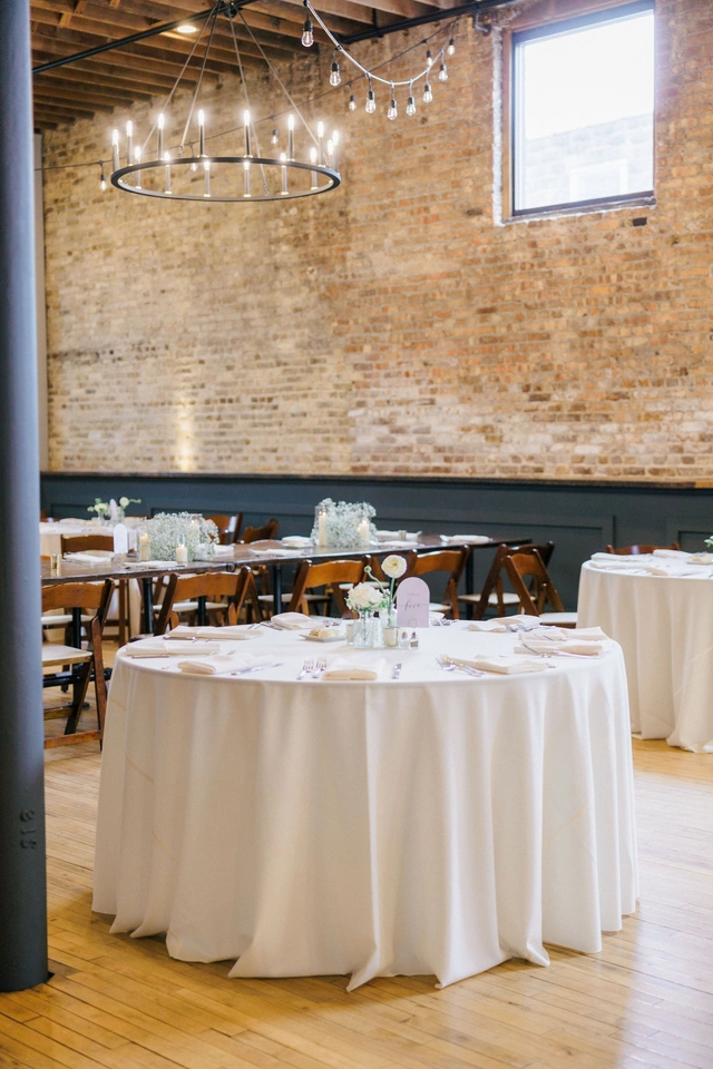 Round wedding reception table with white linens and minimalist centerpiece under string lights at Mercantile Hall