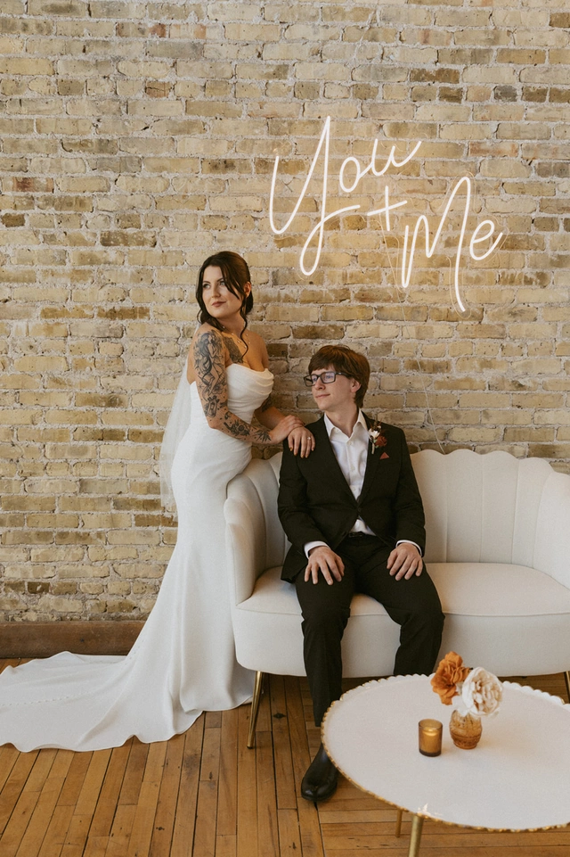 Modern bride and groom pose in front of a brick wall with a glowing "You + Me" neon sign at Mercantile Hall