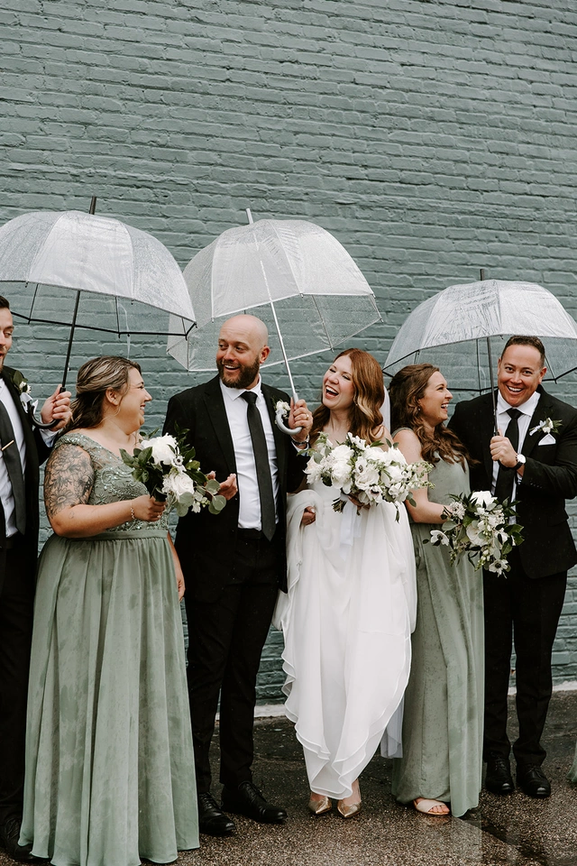 Rainy wedding party photo with clear umbrellas and sage green bridesmaid dresses outside 10 South in Janesville, WI