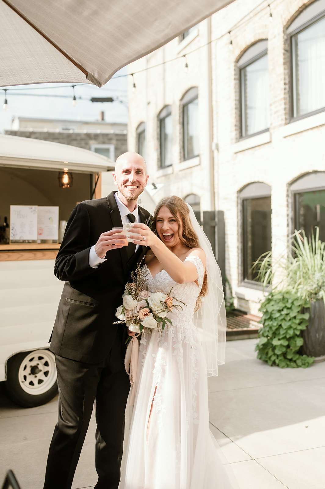 Bride and groom celebrating with drinks in front of a mobile camper bar during outdoor wedding reception