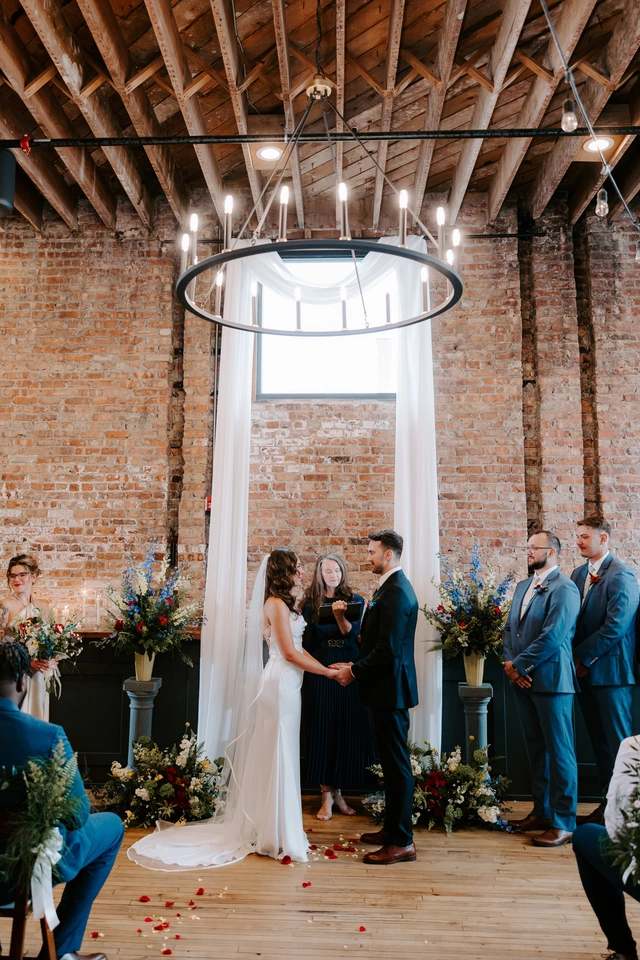 Bride and groom exchanging vows beneath a chandelier and draped window at Mercantile Hall wedding ceremony