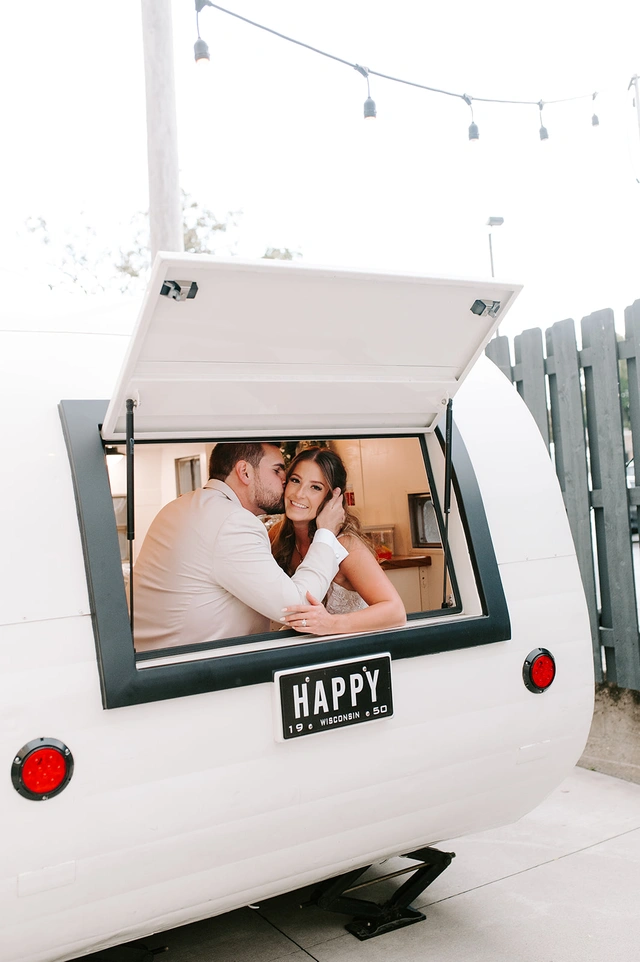Bride and groom cuddling inside the open service window of a vintage white camper bar with a “HAPPY” license plate at Mercantile Hall