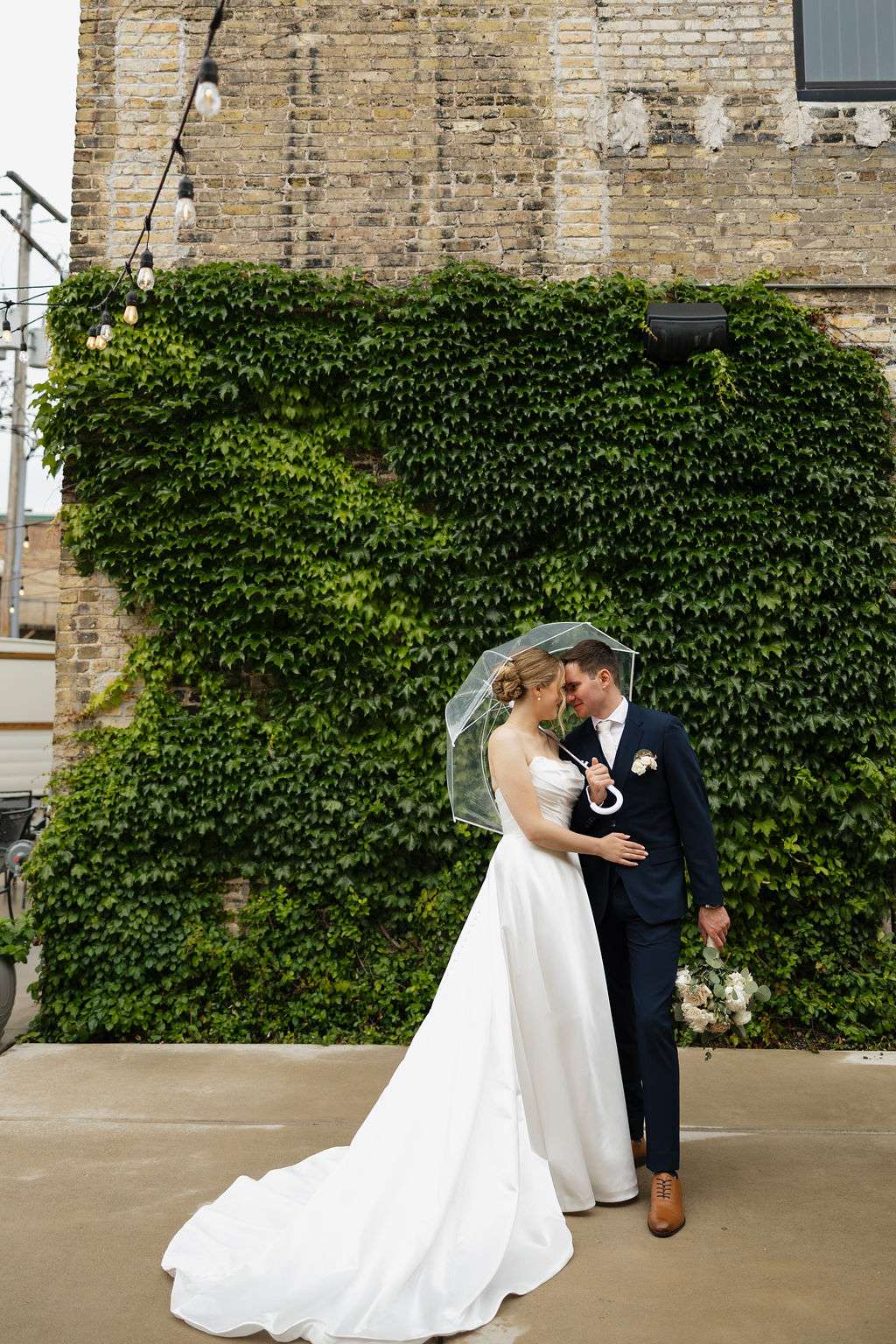 Bride and groom sharing a kiss under a clear umbrella in front of ivy-covered brick wall at Mercantile Hall