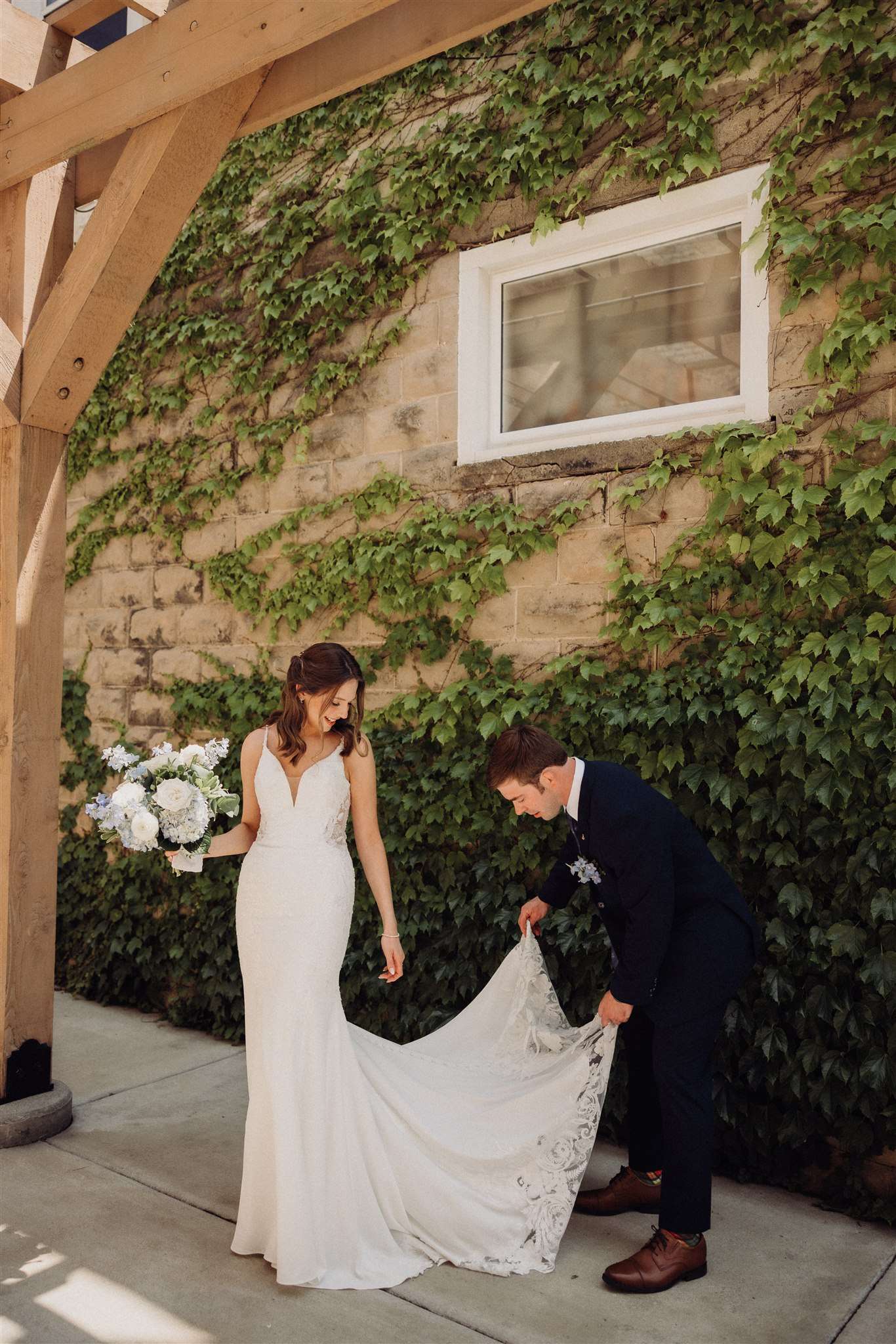 Groom adjusts the train of the bride’s dress as she smiles, standing in front of a vine-covered brick wall at Mercantile Hall.