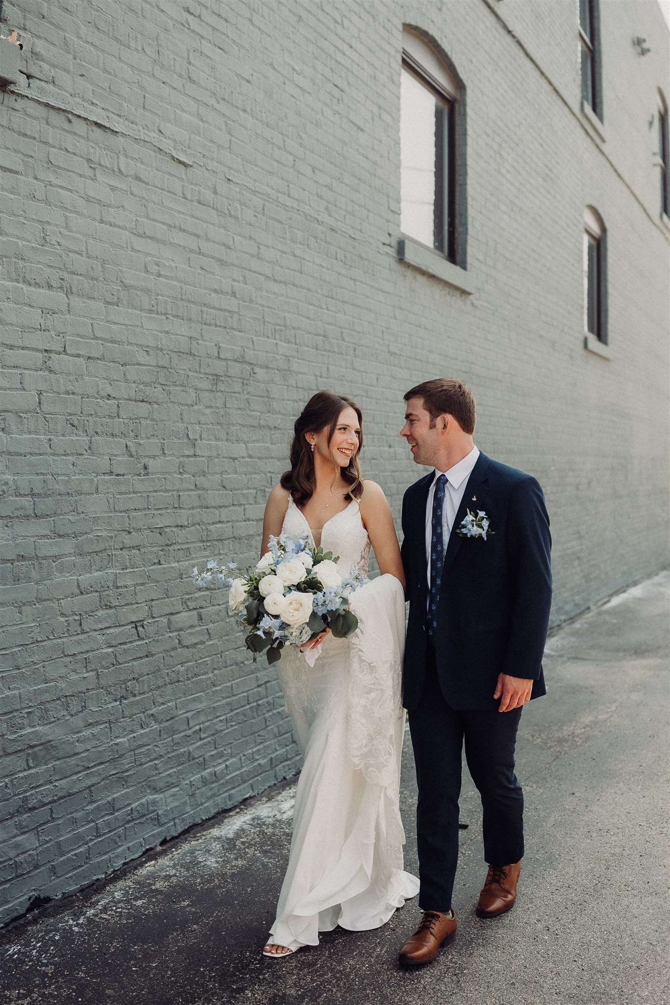 Bride and groom walk together beside a painted brick wall in Burlington, Wisconsin, smiling at each other with a white and blue bouquet in hand.
