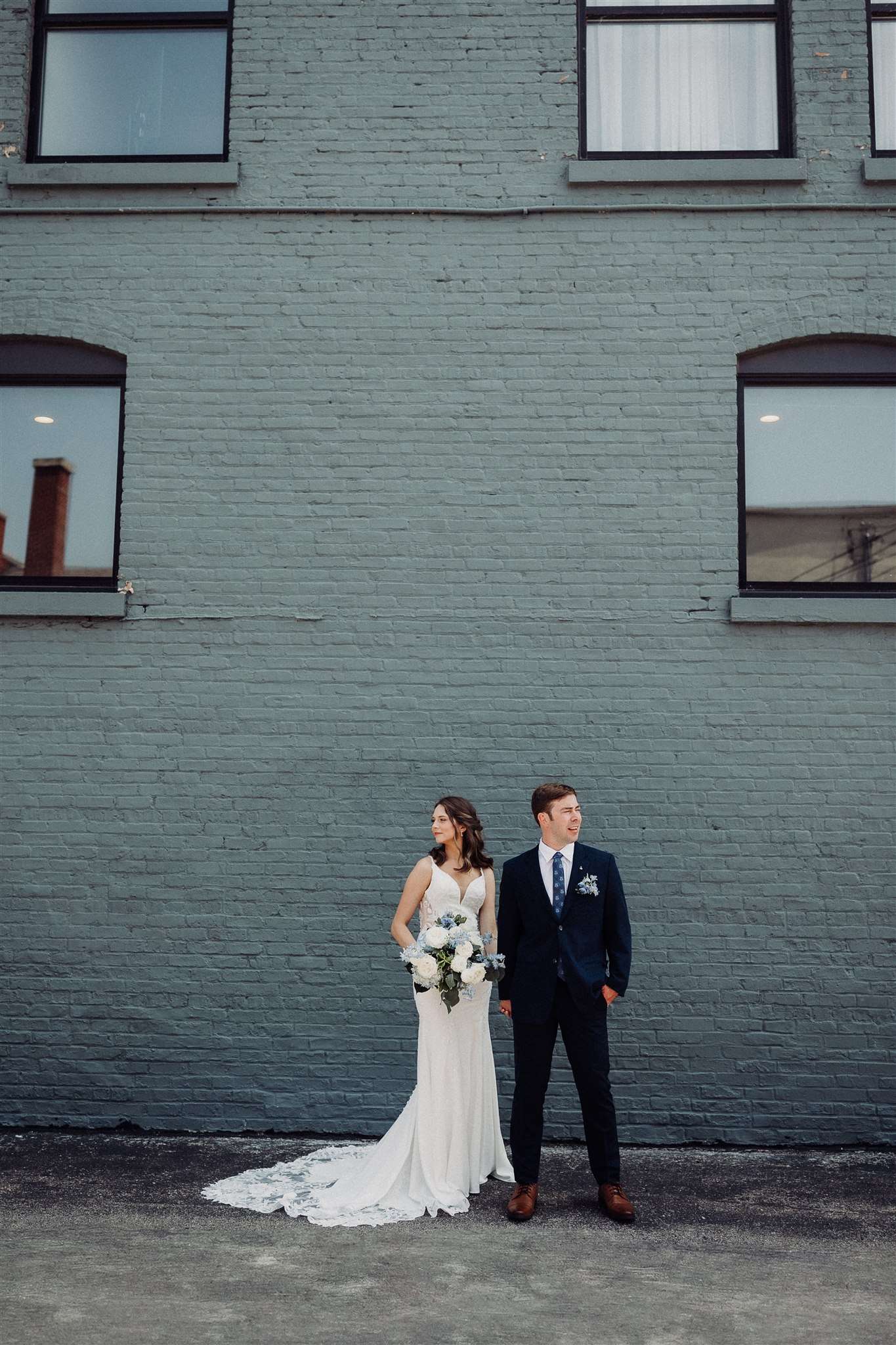 Bride and groom stand side by side against a painted brick wall in Burlington, Wisconsin, each looking in a different direction while holding a white and blue floral bouquet.