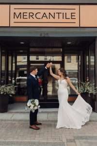 Bride and groom dancing outside Mercantile Hall's entrance in Burlington, Wisconsin, with the bride lifting her train and the groom holding her bouquet of white and blue flowers.