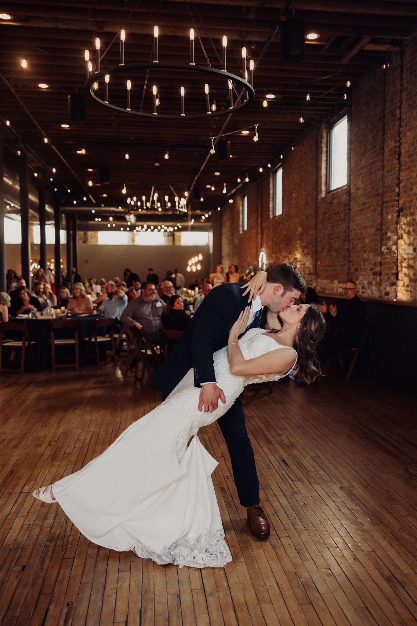 Bride and groom share a dramatic dip and kiss during their first dance in a brick-walled reception hall.