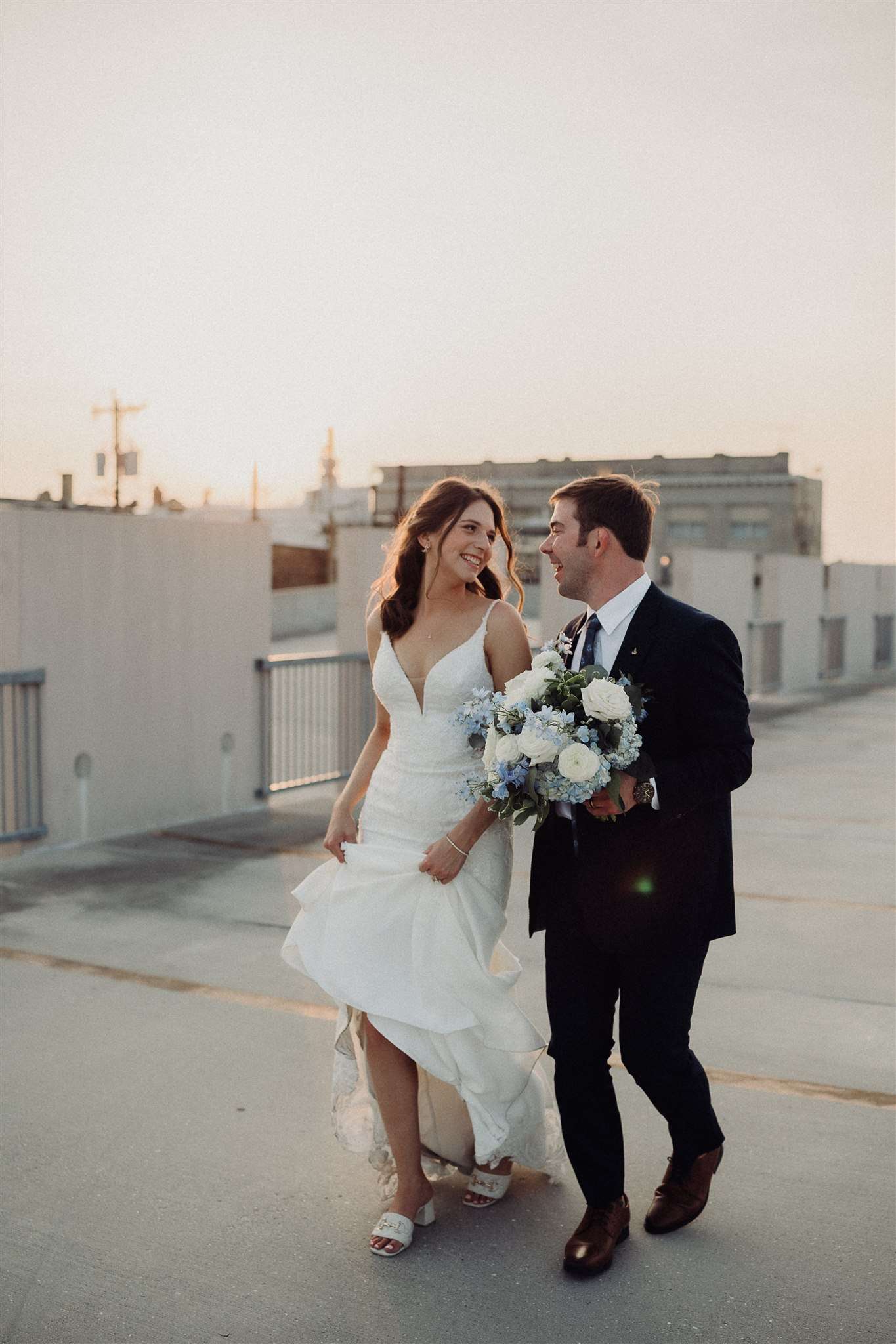 Bride and groom walking hand-in-hand on a rooftop at sunset, smiling and looking at each other.