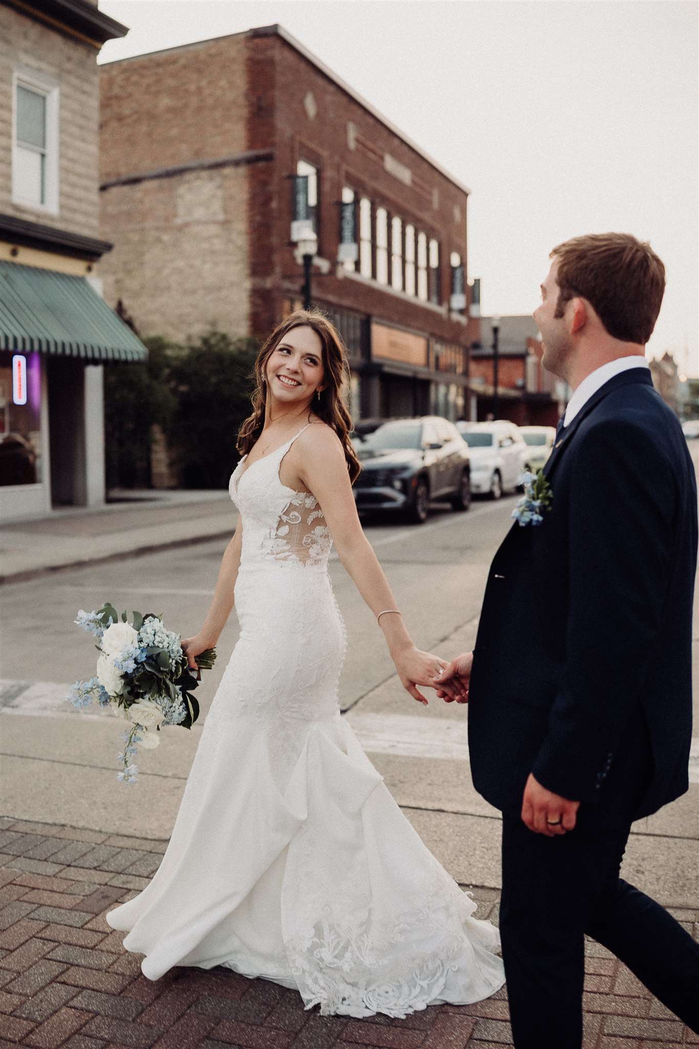 Bride and groom holding hands while walking through a small downtown street, with the bride smiling over her shoulder.