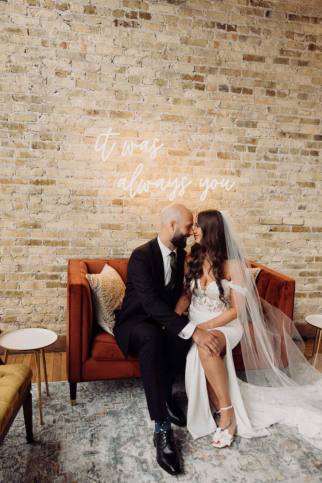 Bride and groom sit close on a velvet loveseat beneath a neon sign that reads "It was always you" at Mercantile Hall