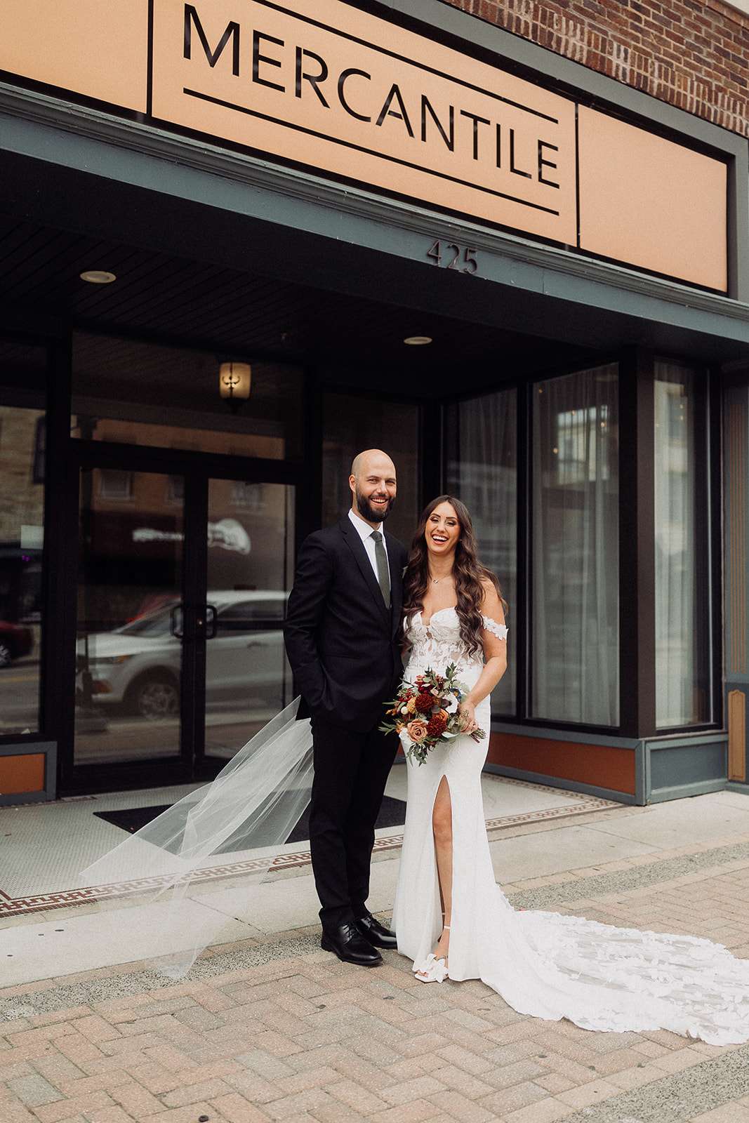 Bride and groom stand smiling outside Mercantile Hall's front entrance under the marquee sign