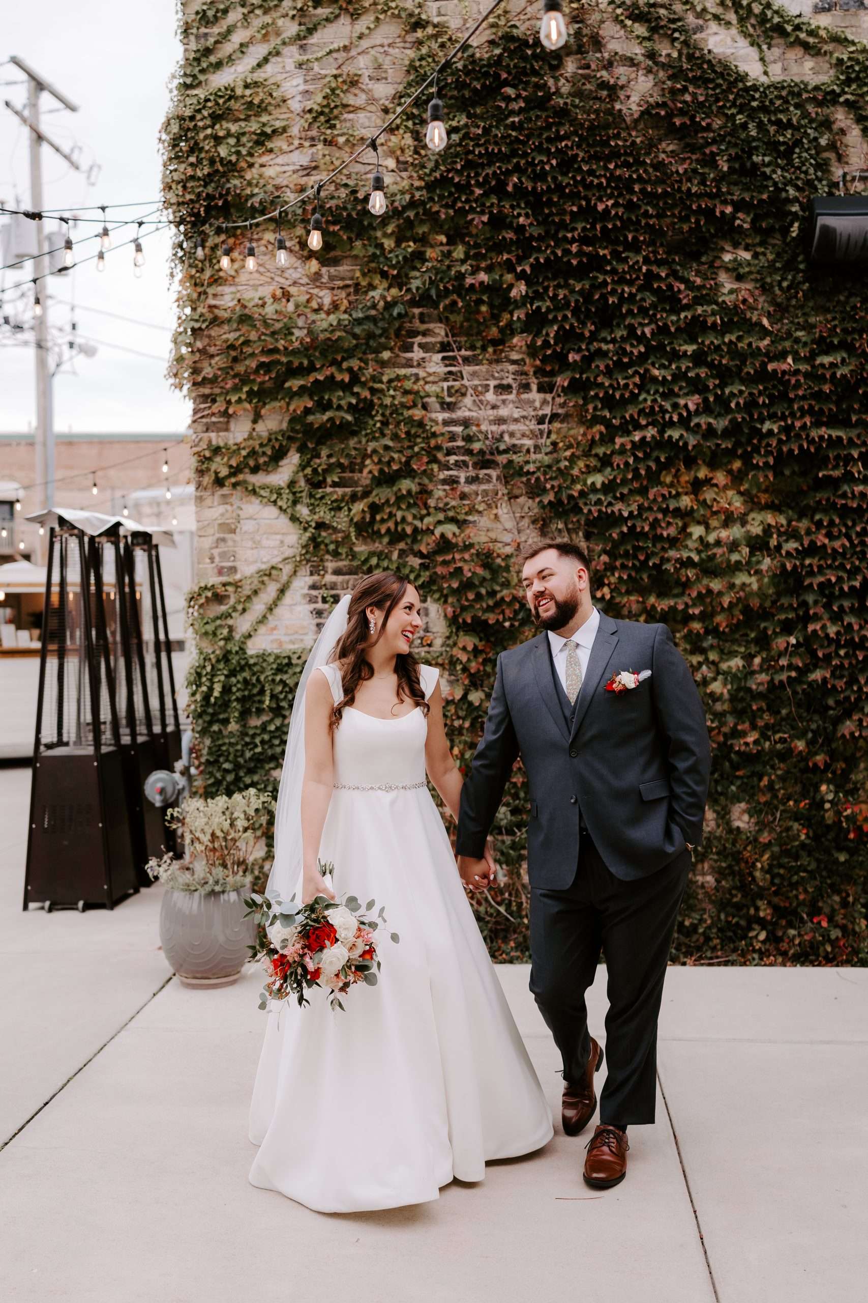 Bride and groom walking hand in hand in front of ivy-covered brick wall at outdoor wedding venue