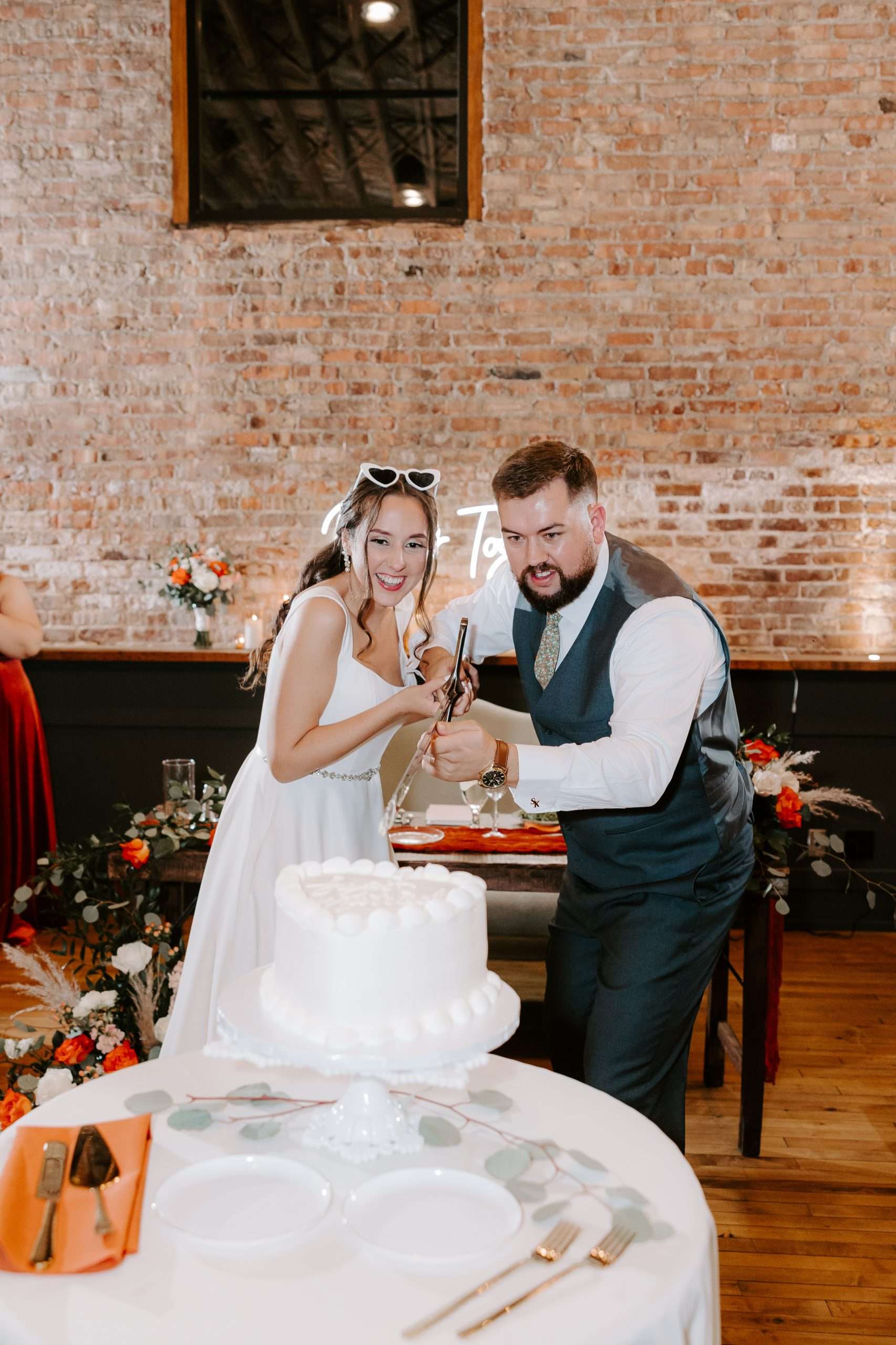 Bride and groom playfully cut their wedding cake in front of an exposed brick wall at their reception