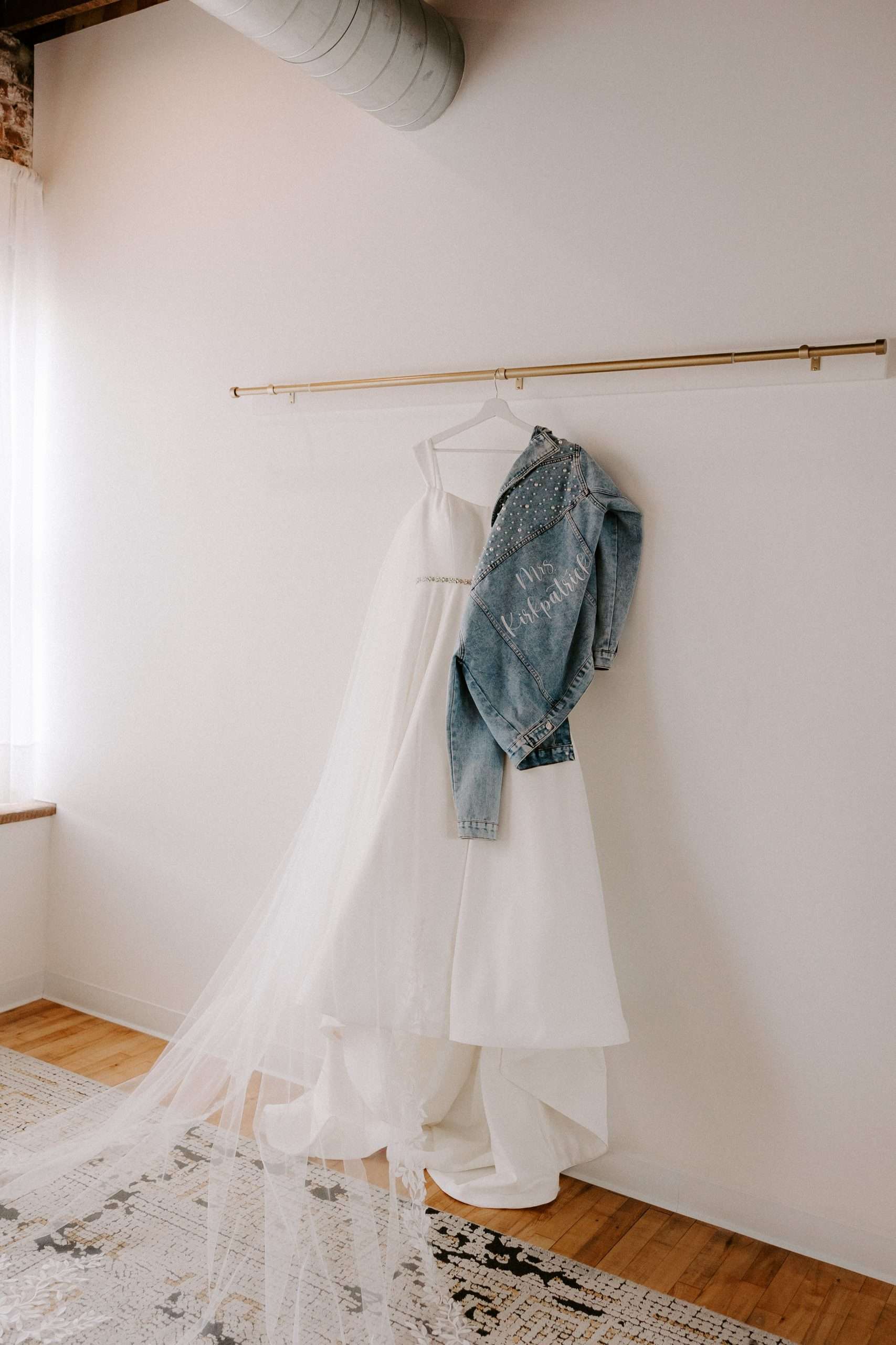 Simple white wedding gown with long veil and embellished denim jacket that reads “Mrs. Krajewski” hanging in a sunlit bridal suite
