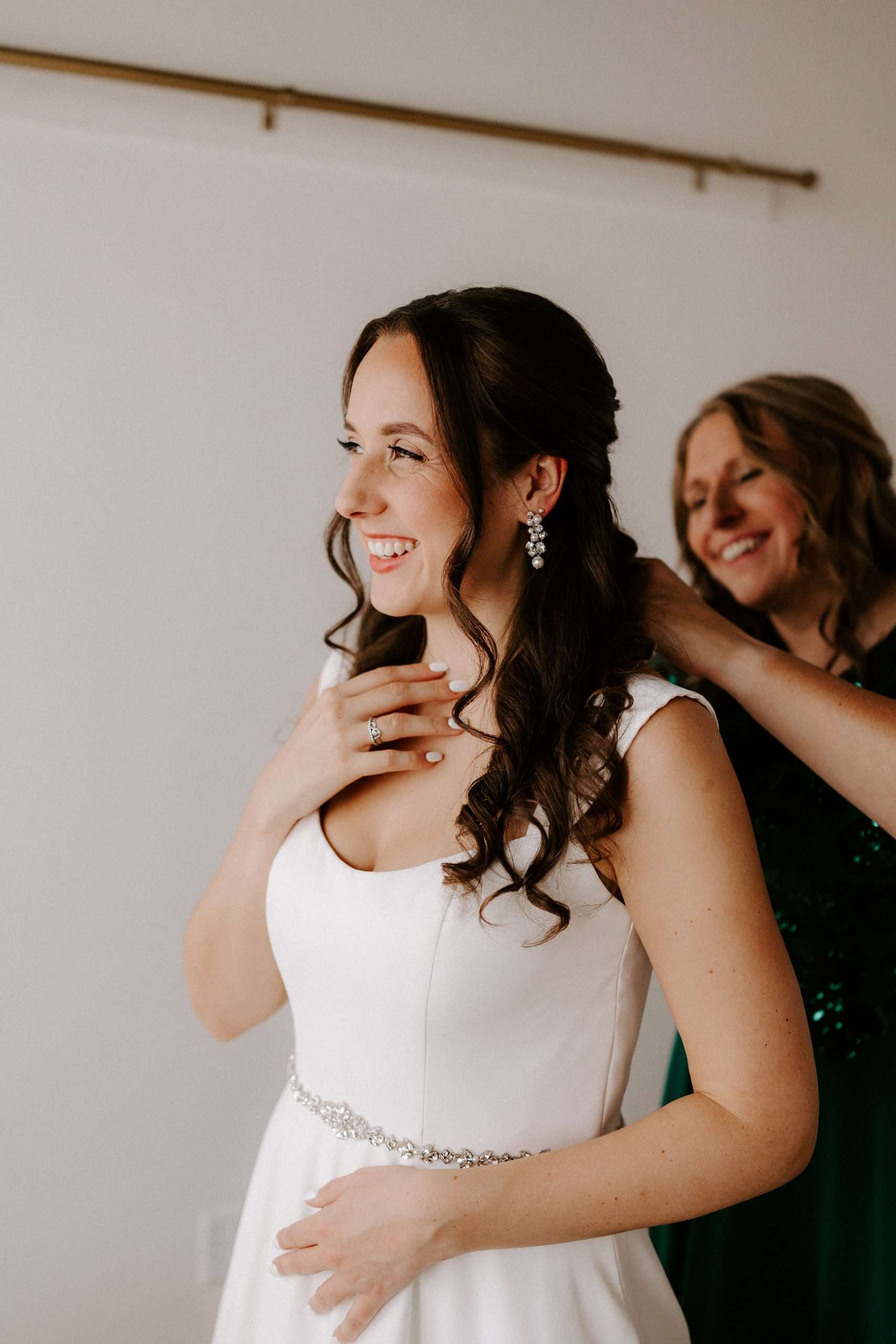 Bride in satin gown smiling as mother helps fasten her necklace before wedding ceremony
