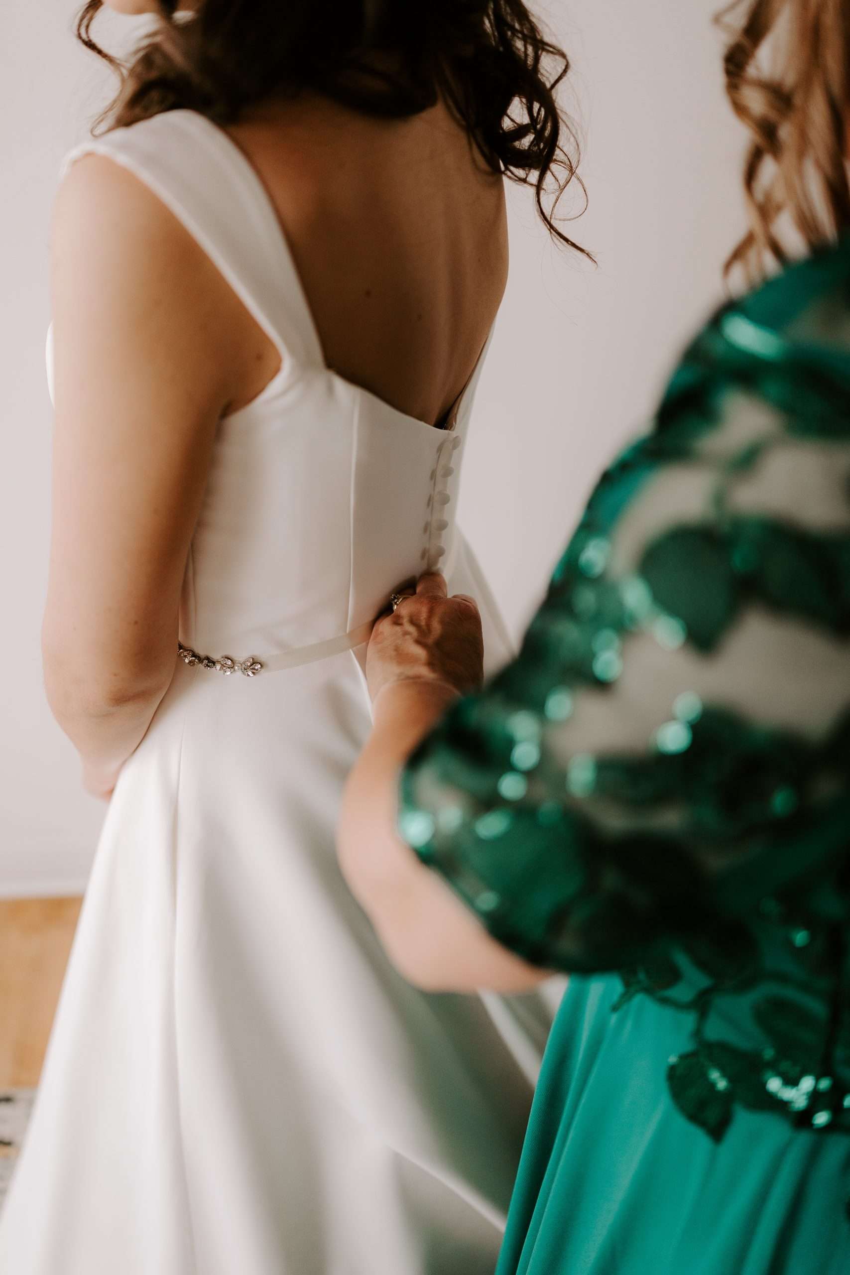 Close-up of bride being zipped into her satin wedding dress by a woman in an emerald green gown