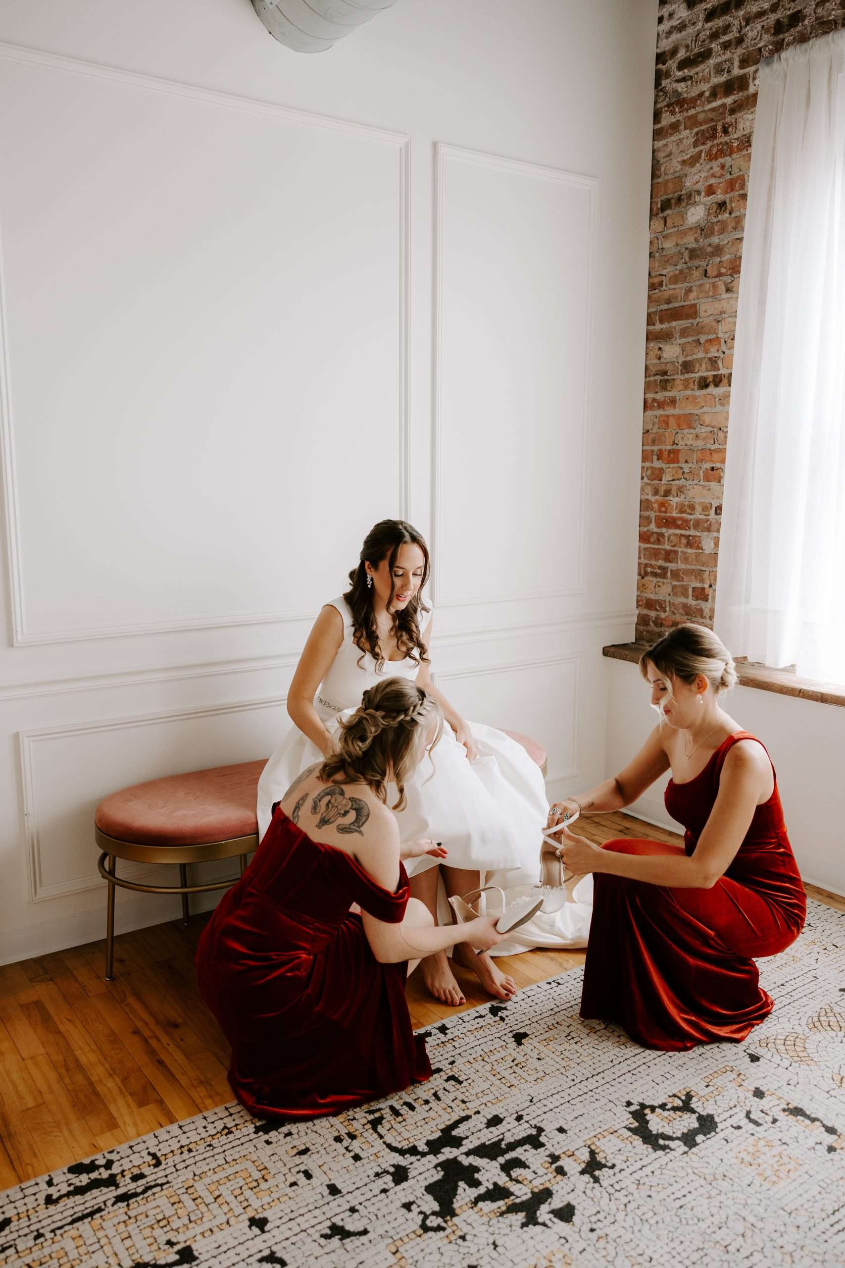 Bridesmaids helping the bride put on her shoes in a softly lit bridal suite