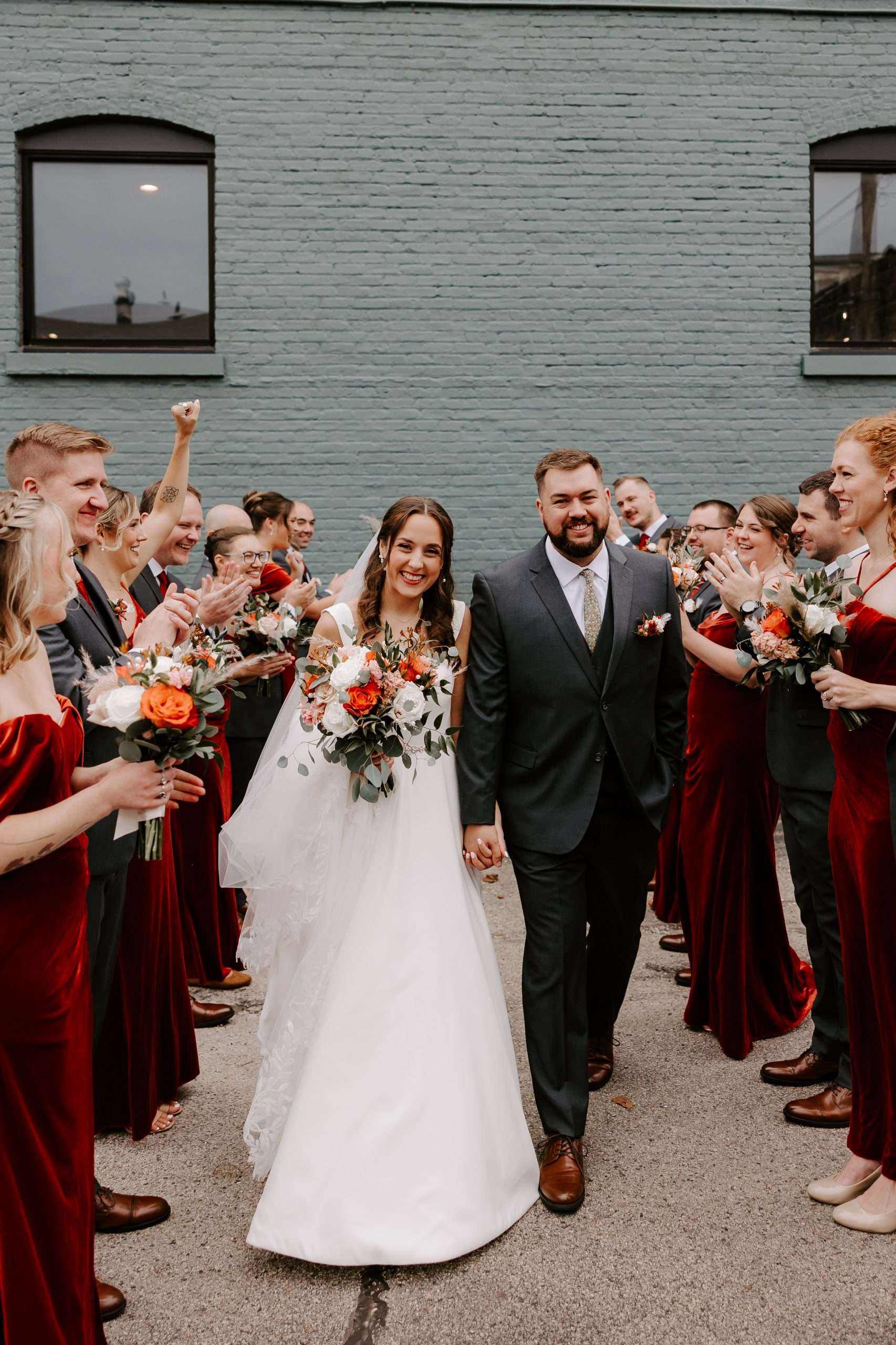 Bride and groom walk hand-in-hand through cheering wedding party outside against a green brick wall