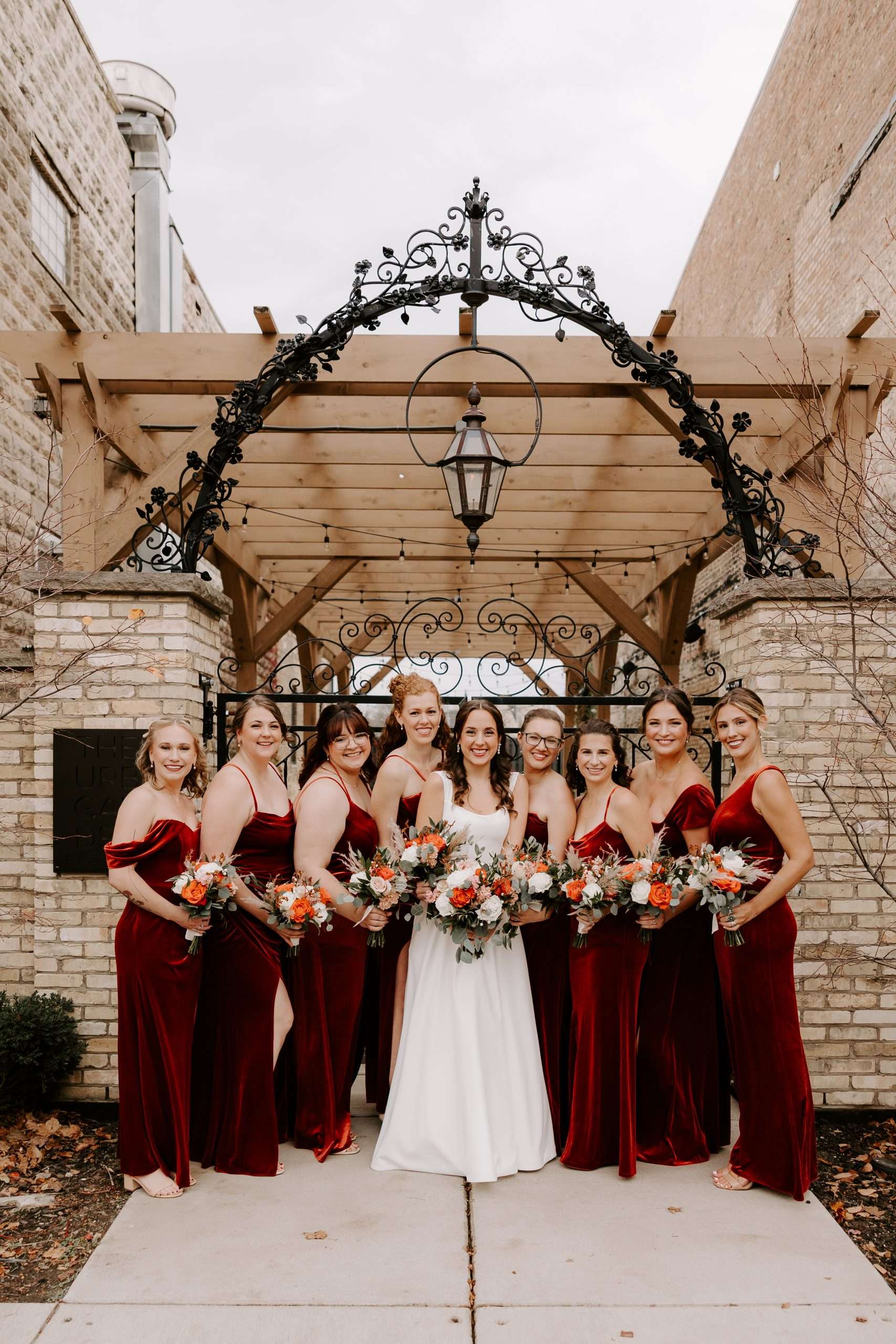 Bride and bridesmaids in burgundy velvet dresses pose under iron pergola at Mercantile Hall in Burlington, WI