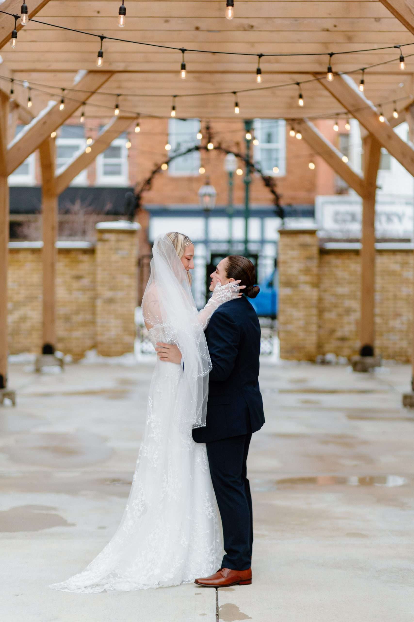Bride and partner sharing a tender moment under string lights on a wooden pergola in an intimate outdoor wedding setting