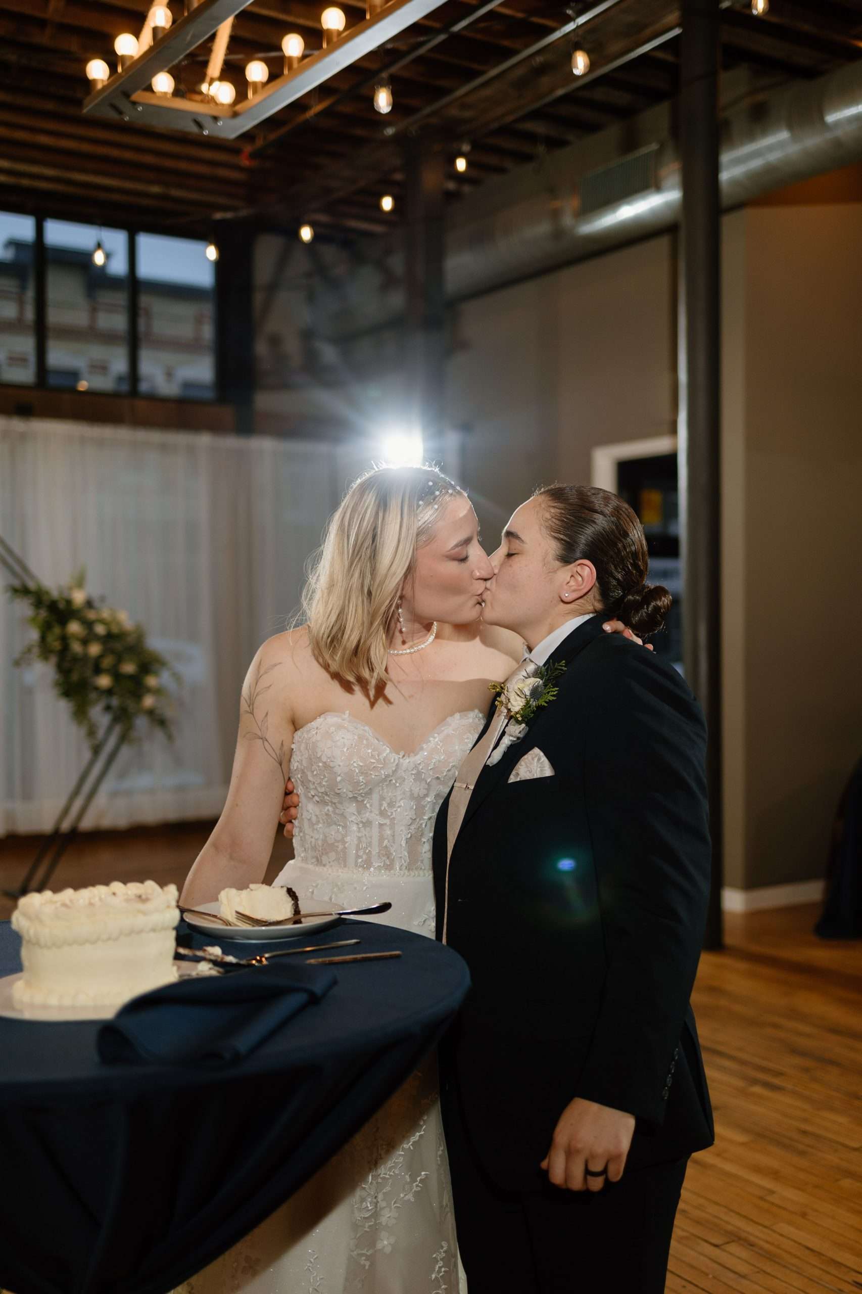 Wedding couple sharing a kiss beside their wedding cake at a warmly lit reception venue with exposed wood beams and string lights