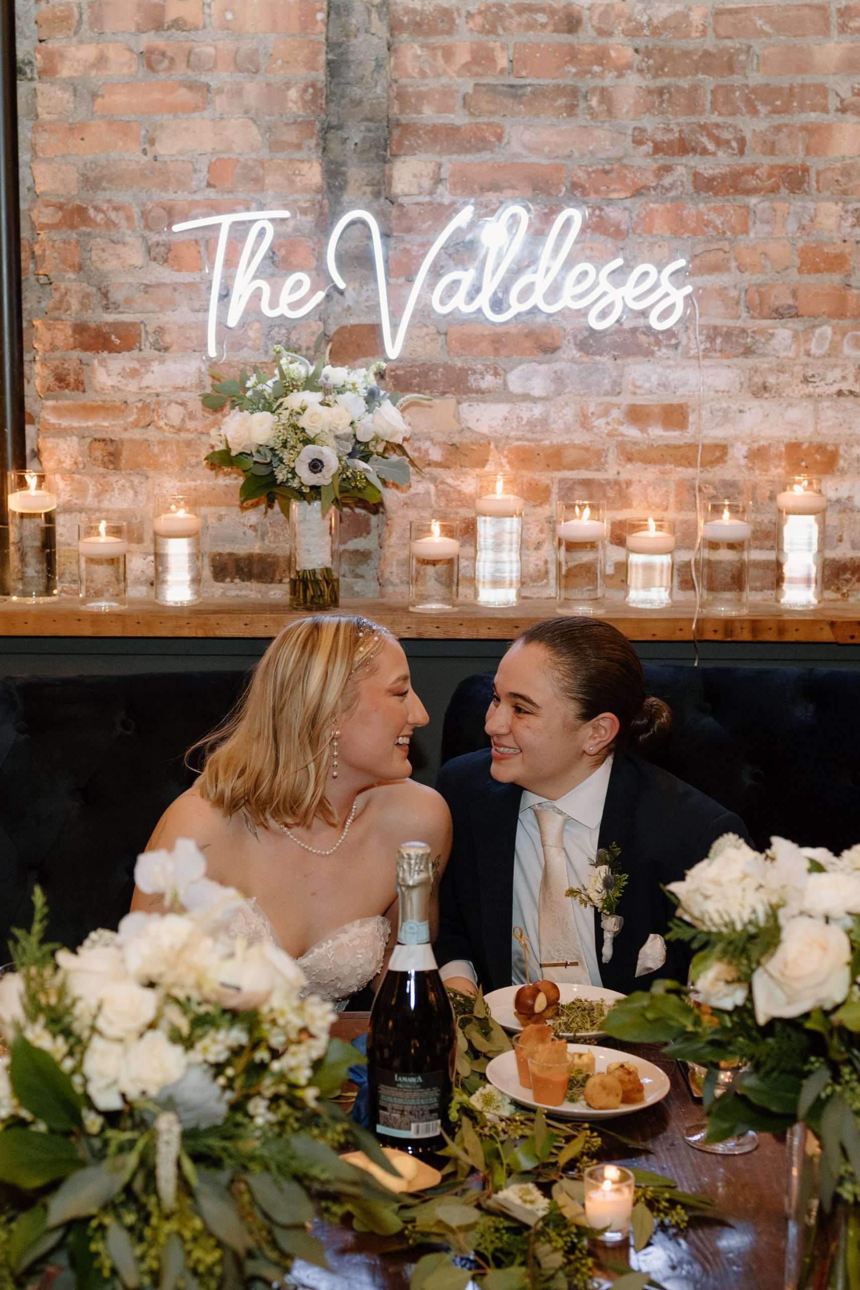 Newlywed couple smiling at each other while seated at their wedding reception table adorned with white flowers, candles, and a personalized neon sign on a brick wall