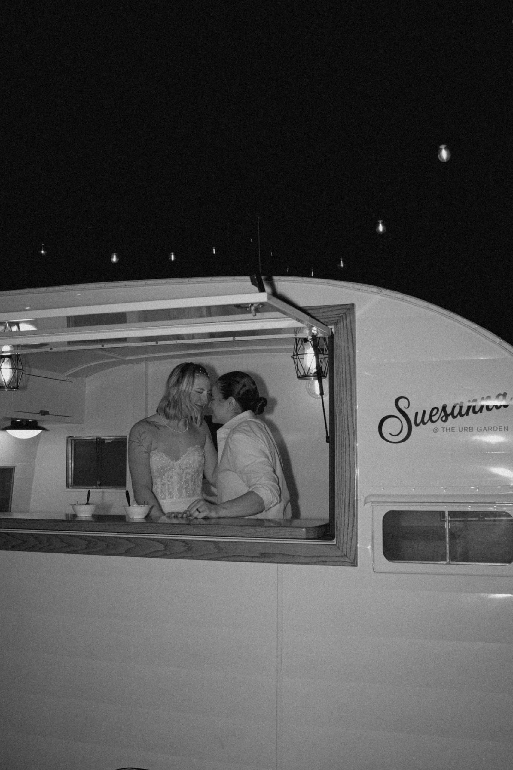 Black and white photo of two brides sharing a tender moment inside a vintage camper bar at night, surrounded by string lights