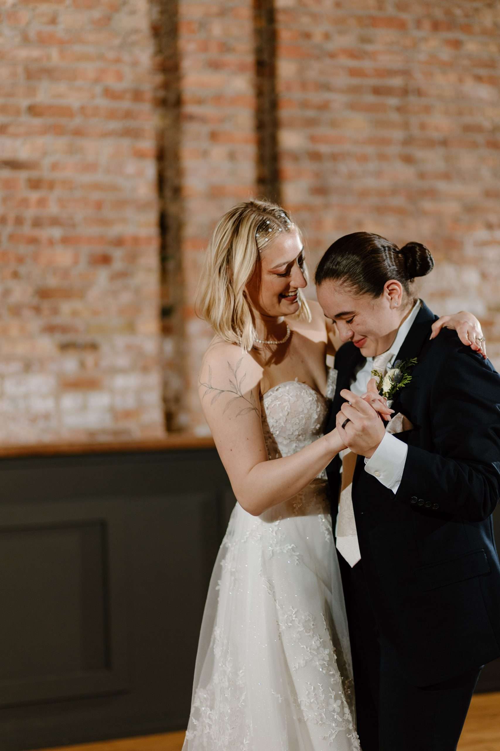 Two brides sharing a joyful first dance in a rustic brick wedding venue, one in a strapless white gown and the other in a black suit with a boutonniere