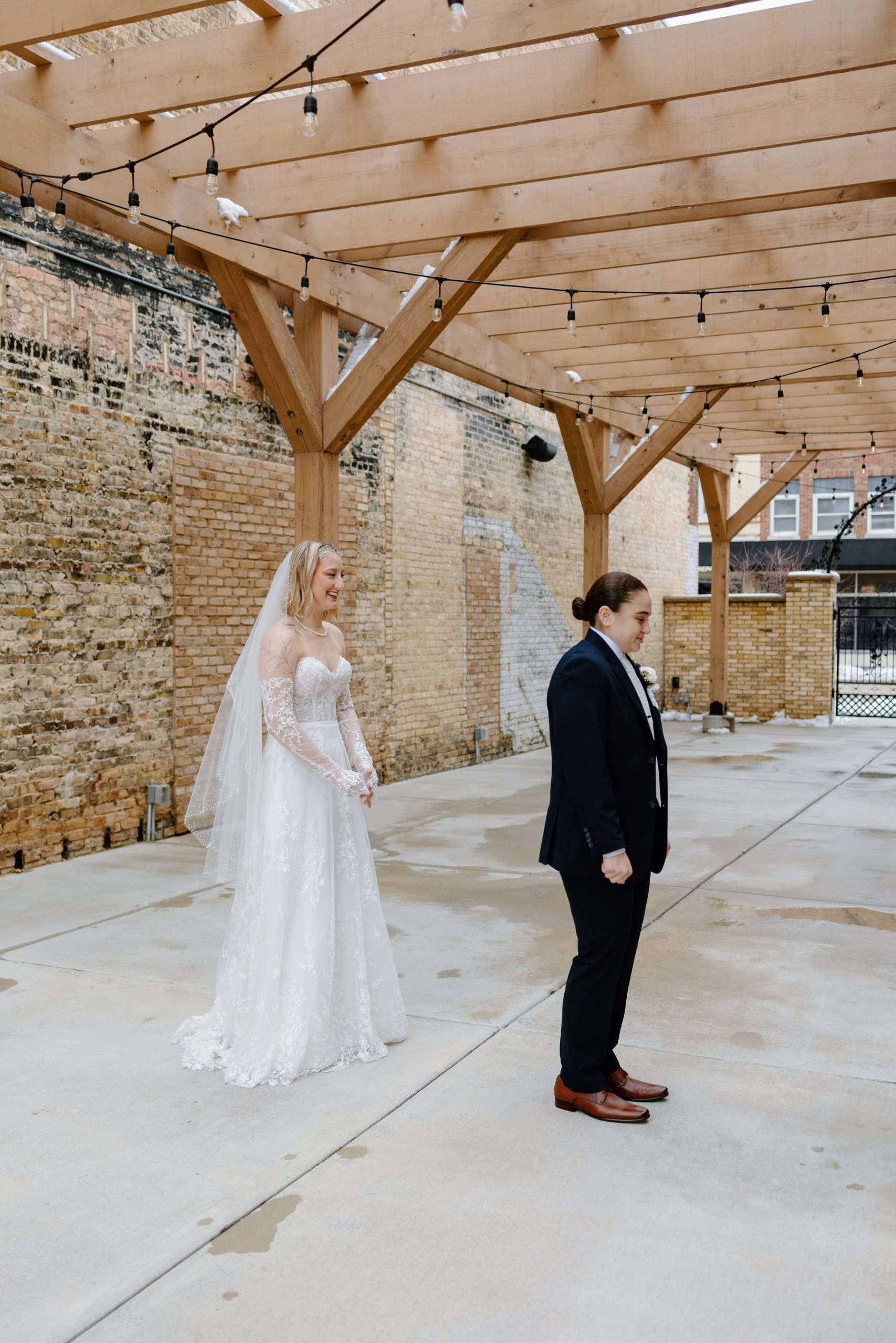 Bride in lace wedding gown standing behind groom in black suit during first look under wooden pergola with string lights and brick walls