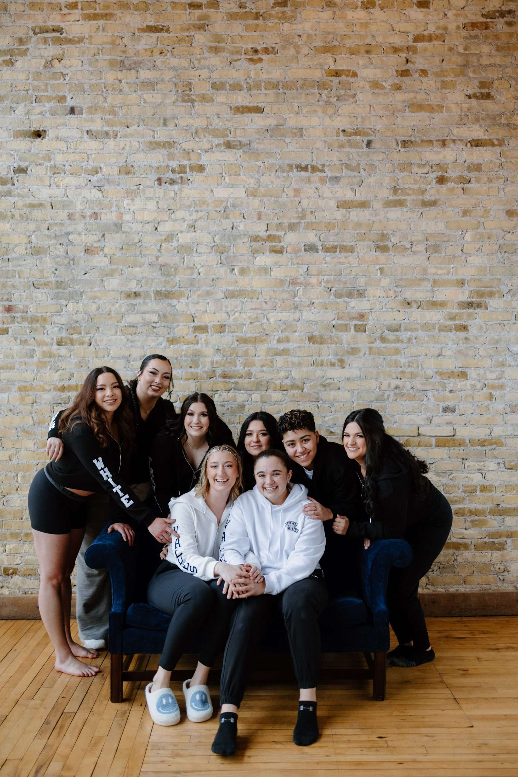 Group portrait of eight friends in casual clothes smiling and posing together indoors against a textured brick wall, with some sitting on a navy blue couch and others standing behind