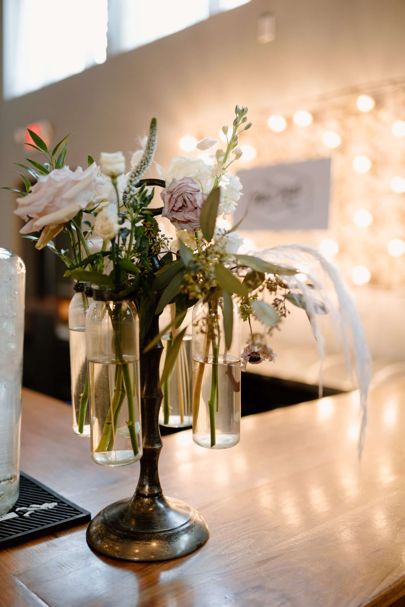 Vintage-style metal floral centerpiece with hanging glass vases holding white and pale pink flowers on wooden table, warm string light background
