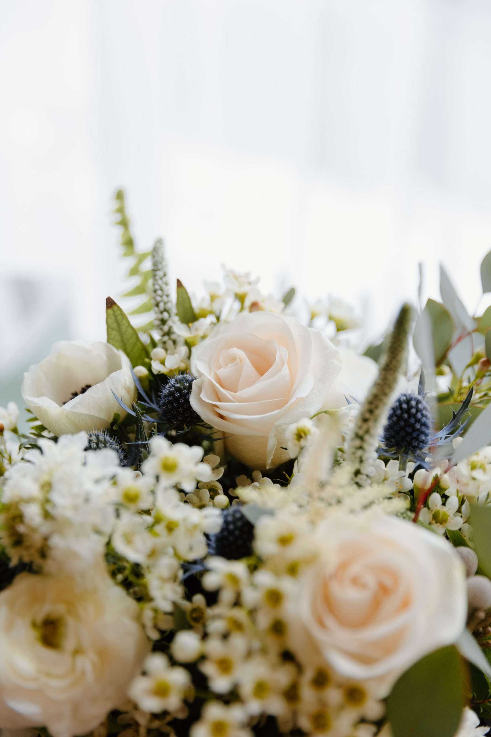 Close-up of wedding bouquet with pale peach roses, white flowers, blue thistles, and greenery against soft light background