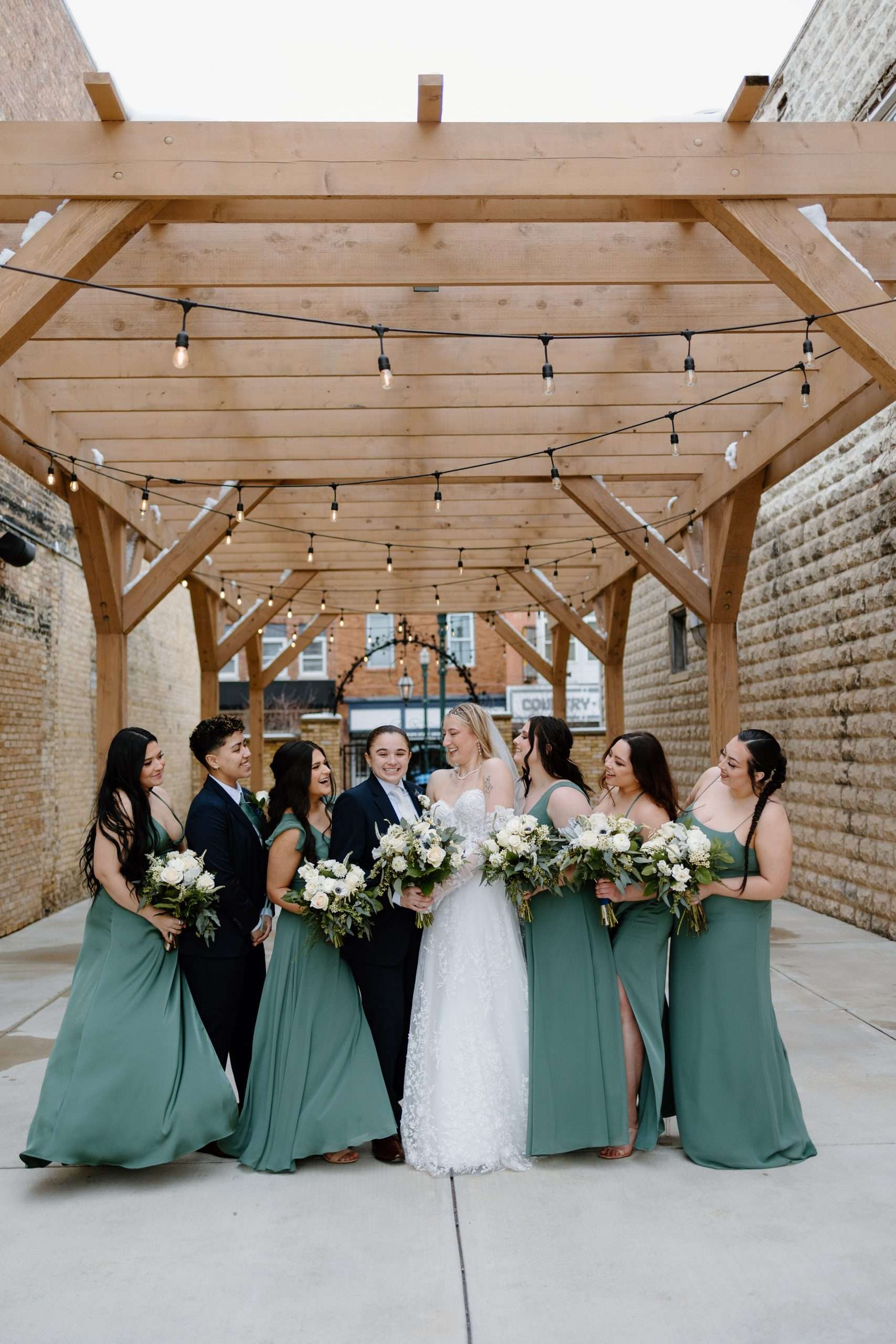Bride and wedding party smiling under wooden pergola with string lights, bridesmaids in sage green dresses, two grooms in black suits, all holding bouquets of white and green flowers, outdoor urban setting