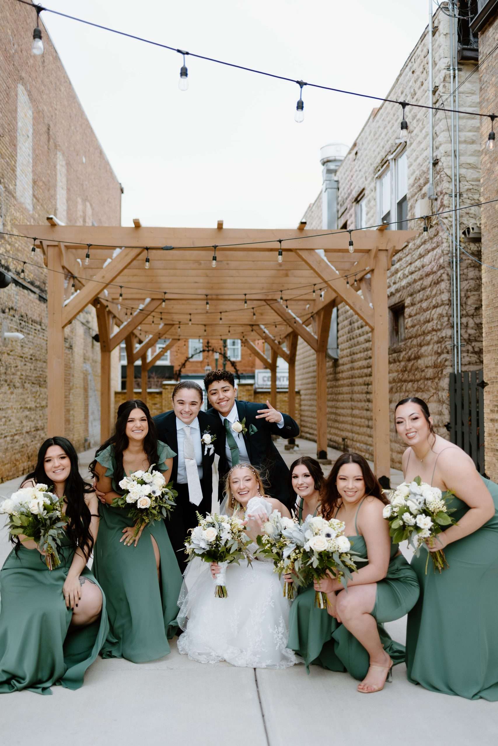 Bride with bridesmaids in sage green dresses and two grooms in black suits posing under wooden pergola with string lights, holding bouquets of white flowers and greenery, urban courtyard setting