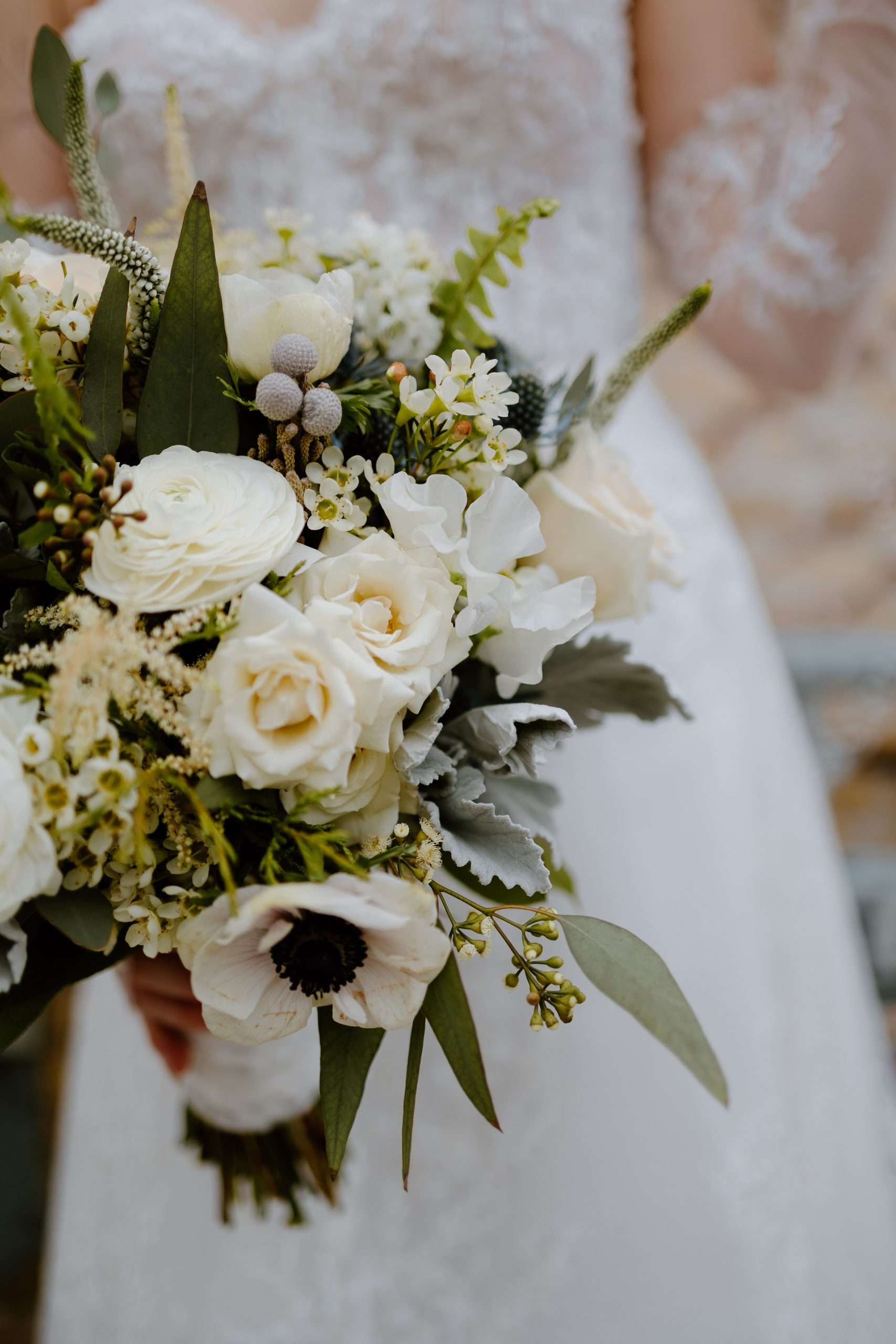 Close-up of bridal bouquet with white roses, anemones, ranunculus, wax flowers, eucalyptus leaves, and dusty miller greenery held by bride in lace gown