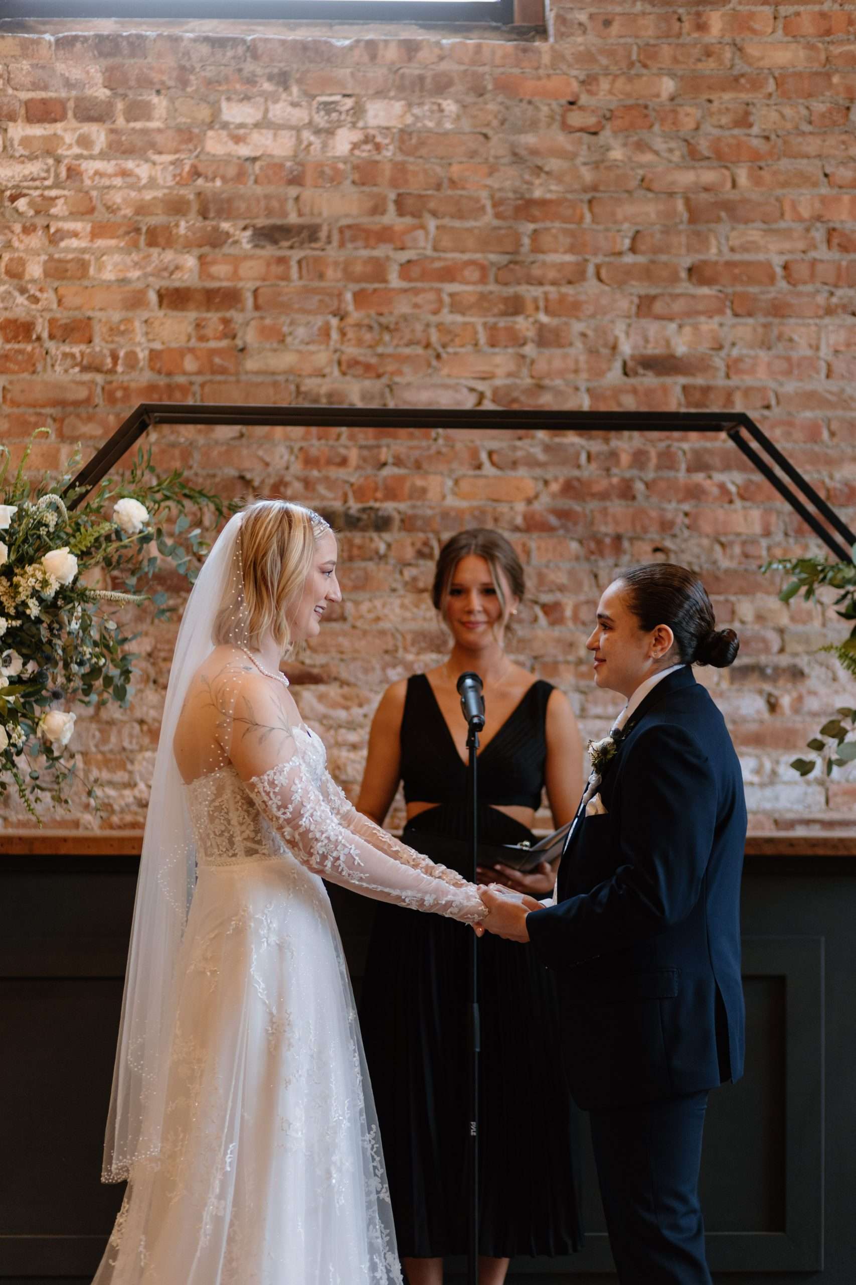 Bride and groom holding hands and smiling during indoor wedding ceremony with officiant and rustic brick wall background