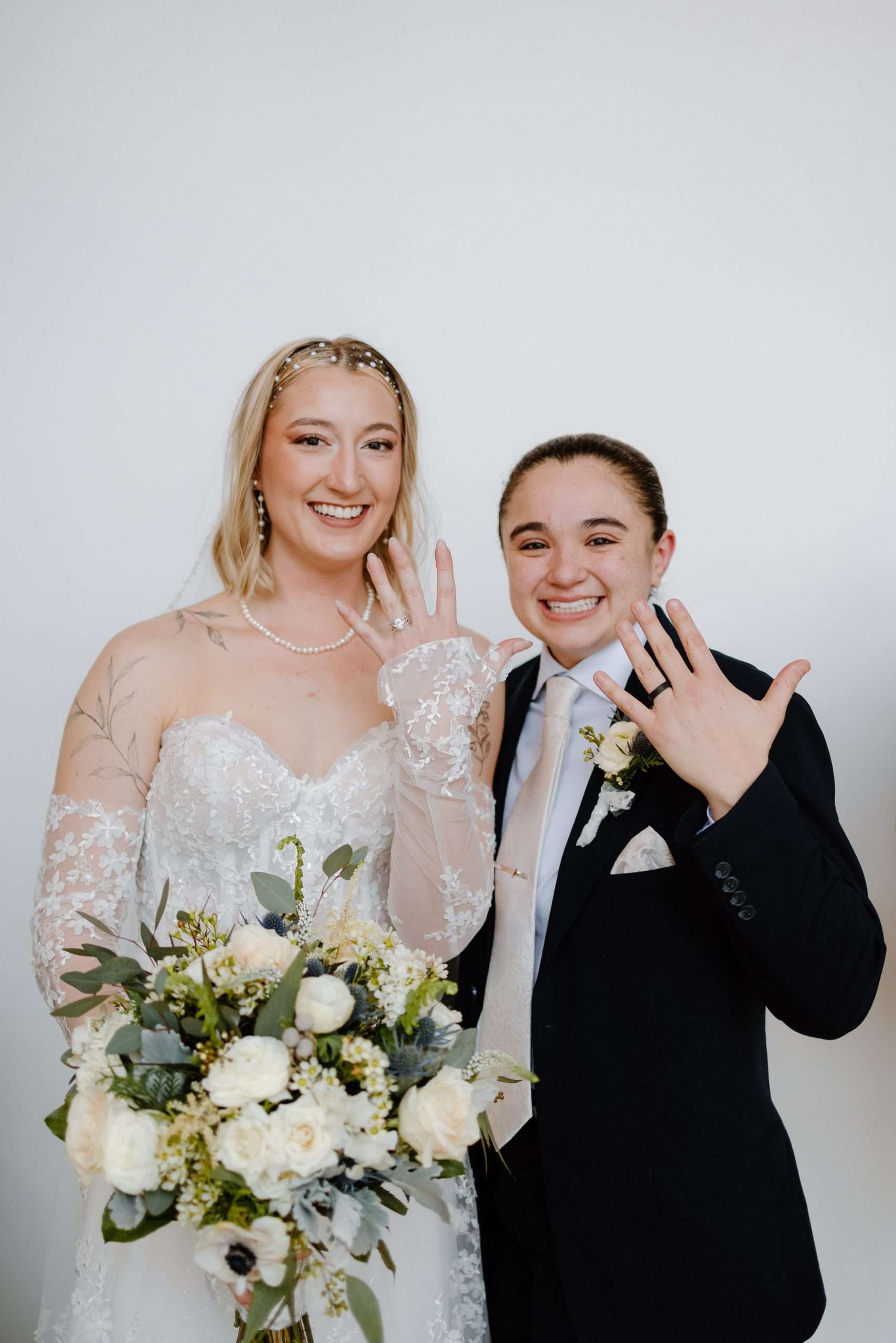Smiling newlywed couple showing wedding rings, bride holding bouquet of white flowers and greenery, groom in black suit with boutonniere, clean white background