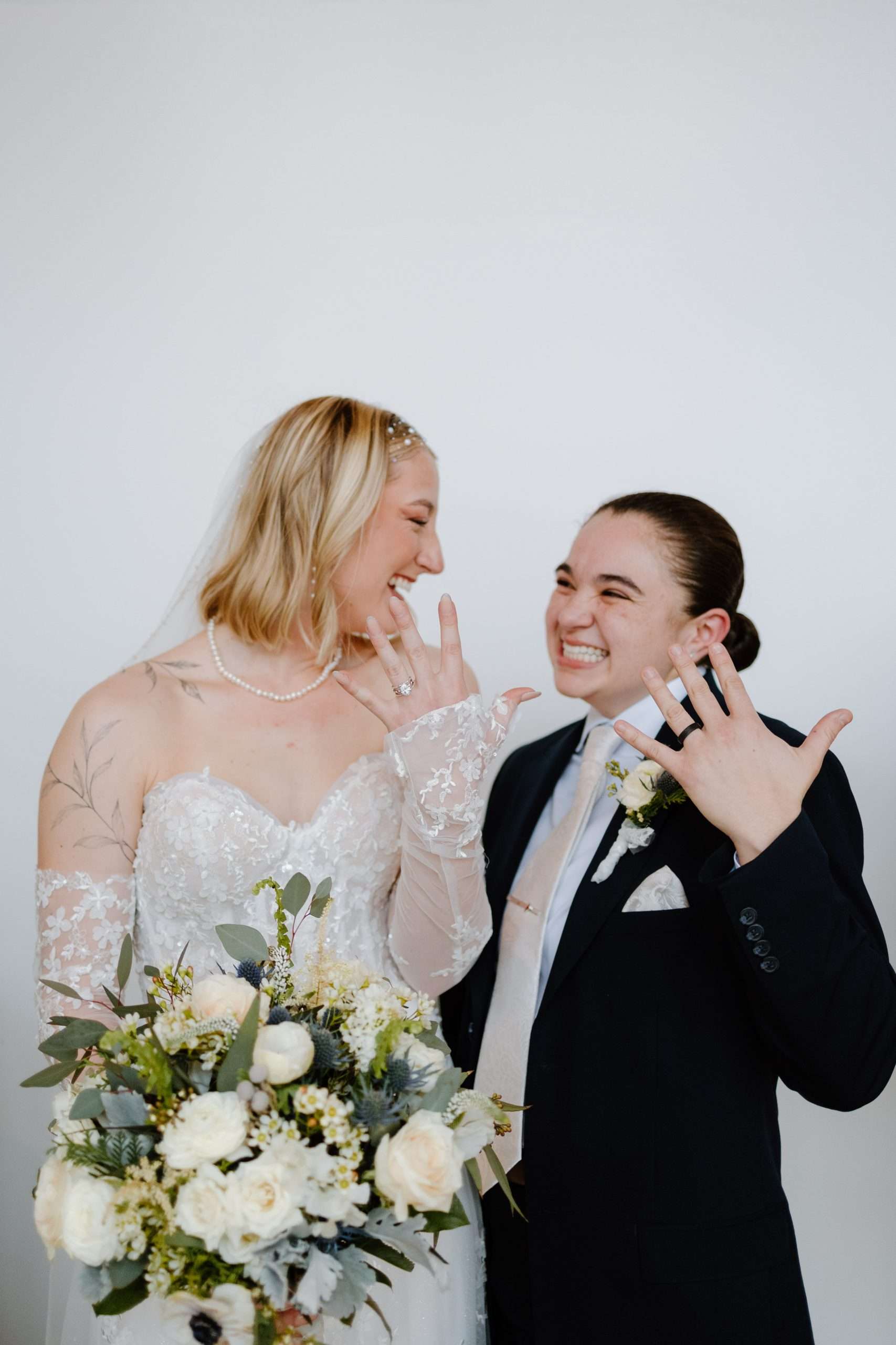 Joyful newlywed couple showing wedding rings with big smiles, bride holding bouquet of white and green flowers, both dressed in elegant wedding attire