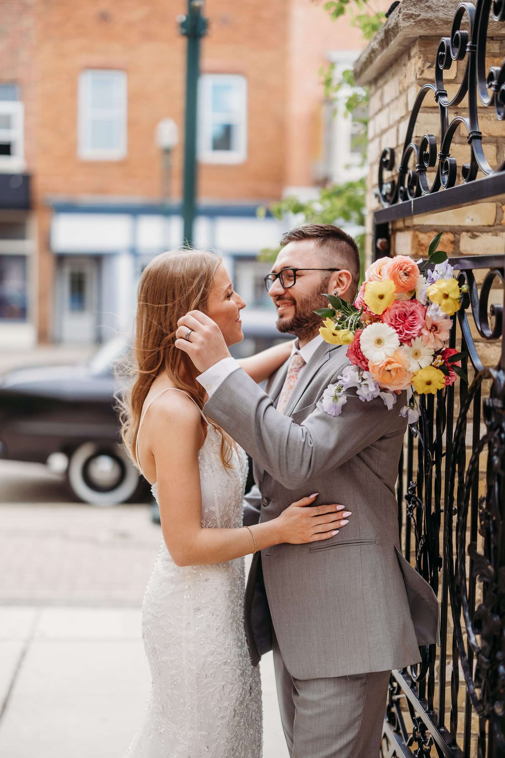 Bride and groom sharing a joyful moment by an ornate iron gate with a colorful bouquet on his shoulder