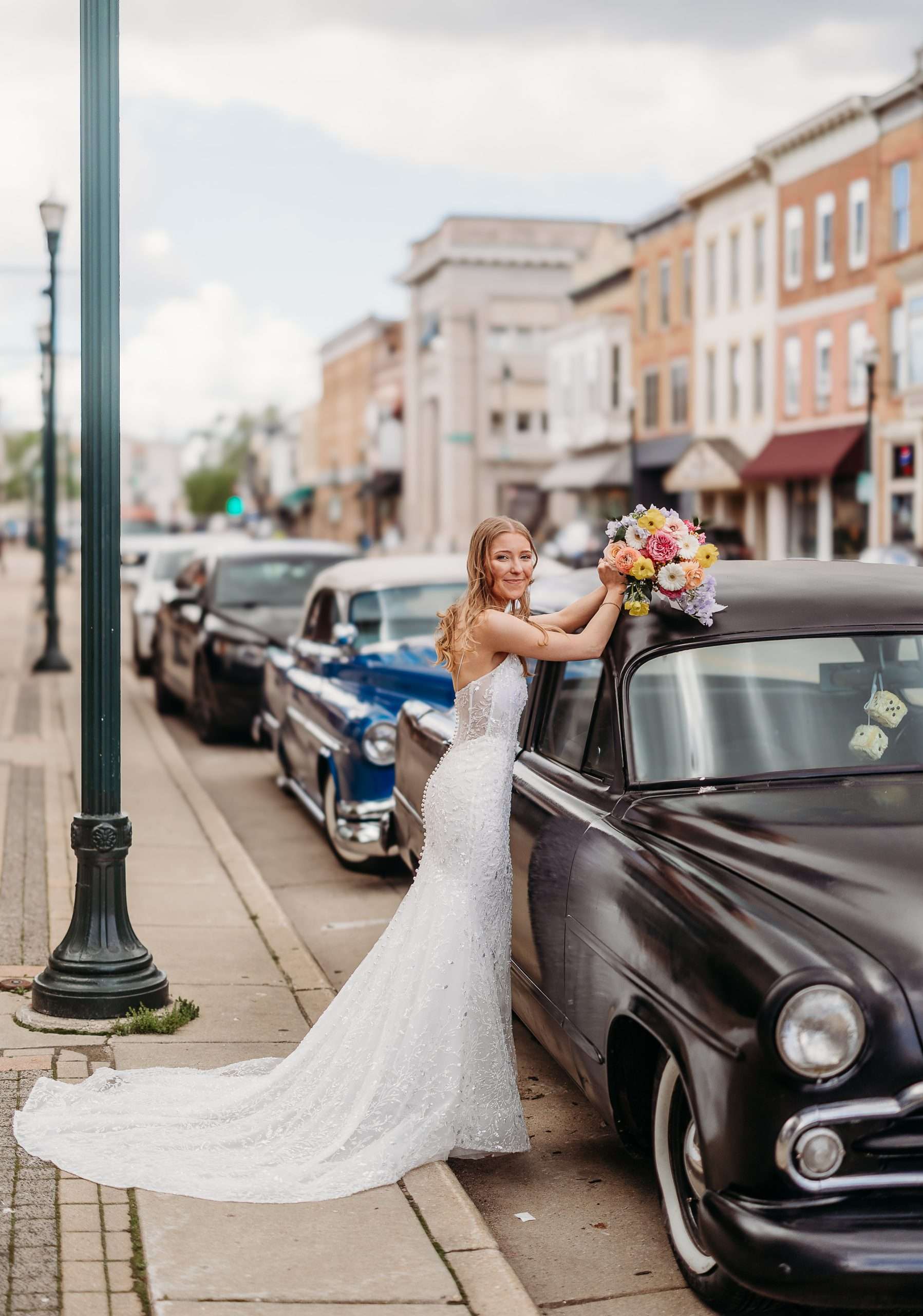 Bride in a beaded gown poses with bouquet on a vintage car in downtown Burlington, Wisconsin
