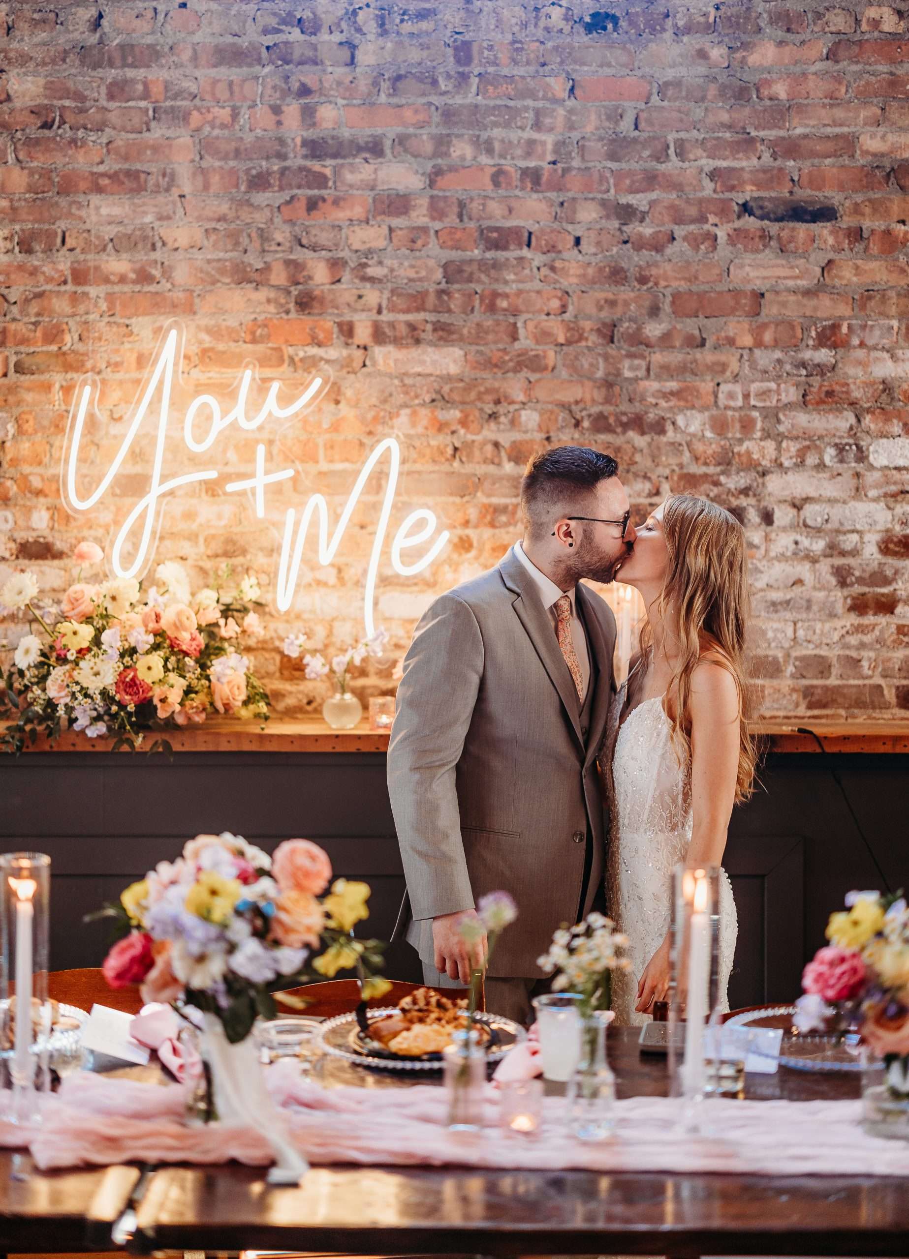 Bride and groom kissing in front of illuminated "You + Me" sign at Mercantile Hall wedding reception with floral arrangements and candlelit tables