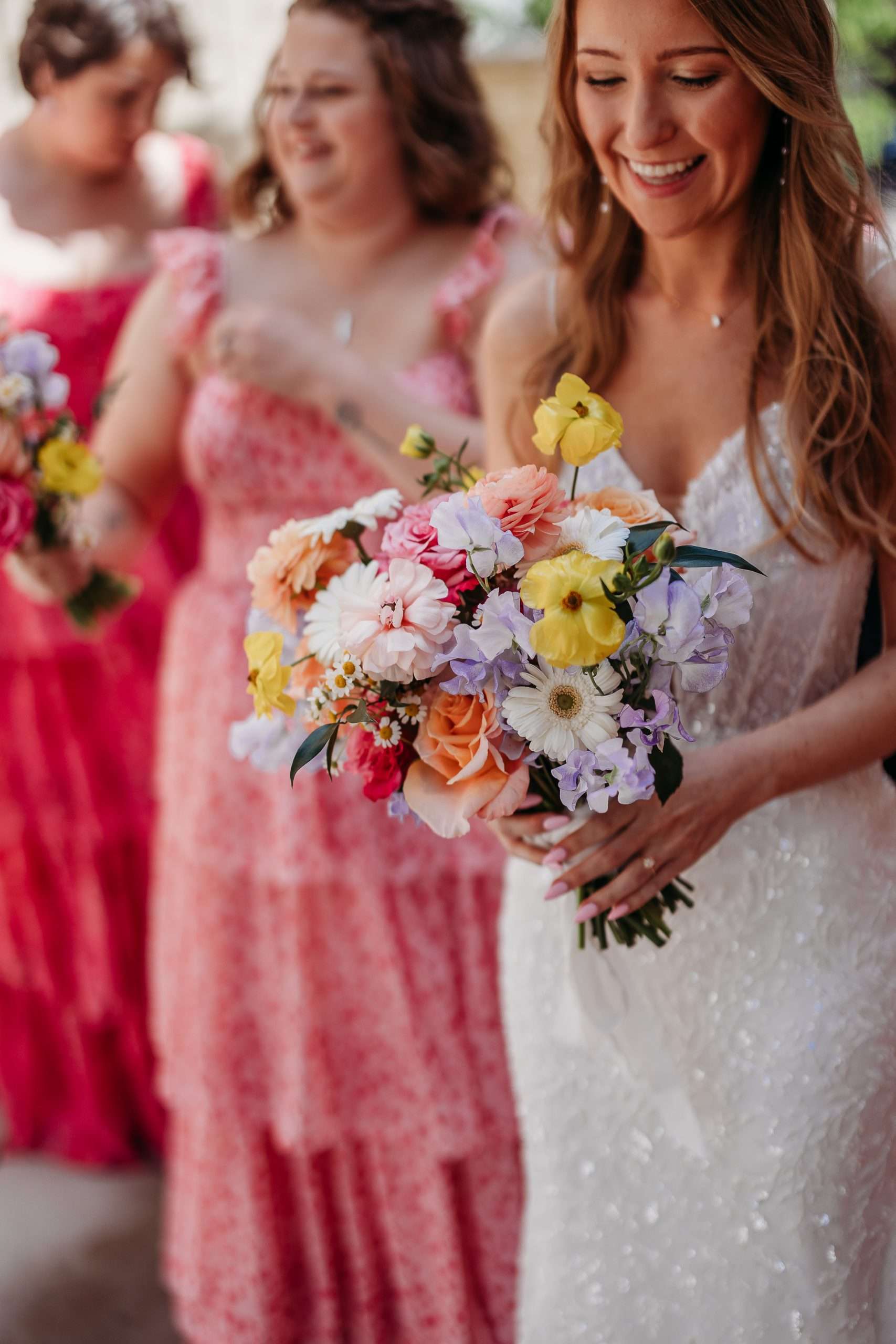 Bride in sequin gown holds colorful wildflower bouquet with bridesmaids in red and pink floral dresses