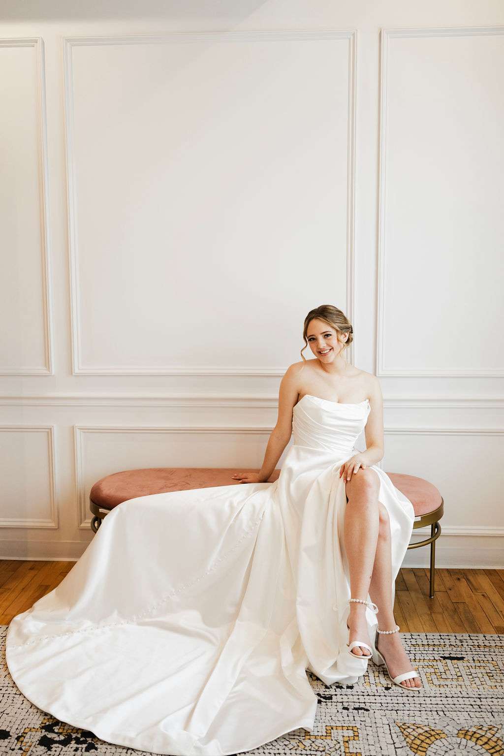Bride in strapless satin gown with high slit seated on pink bench at Bon Bon Belle Bridal