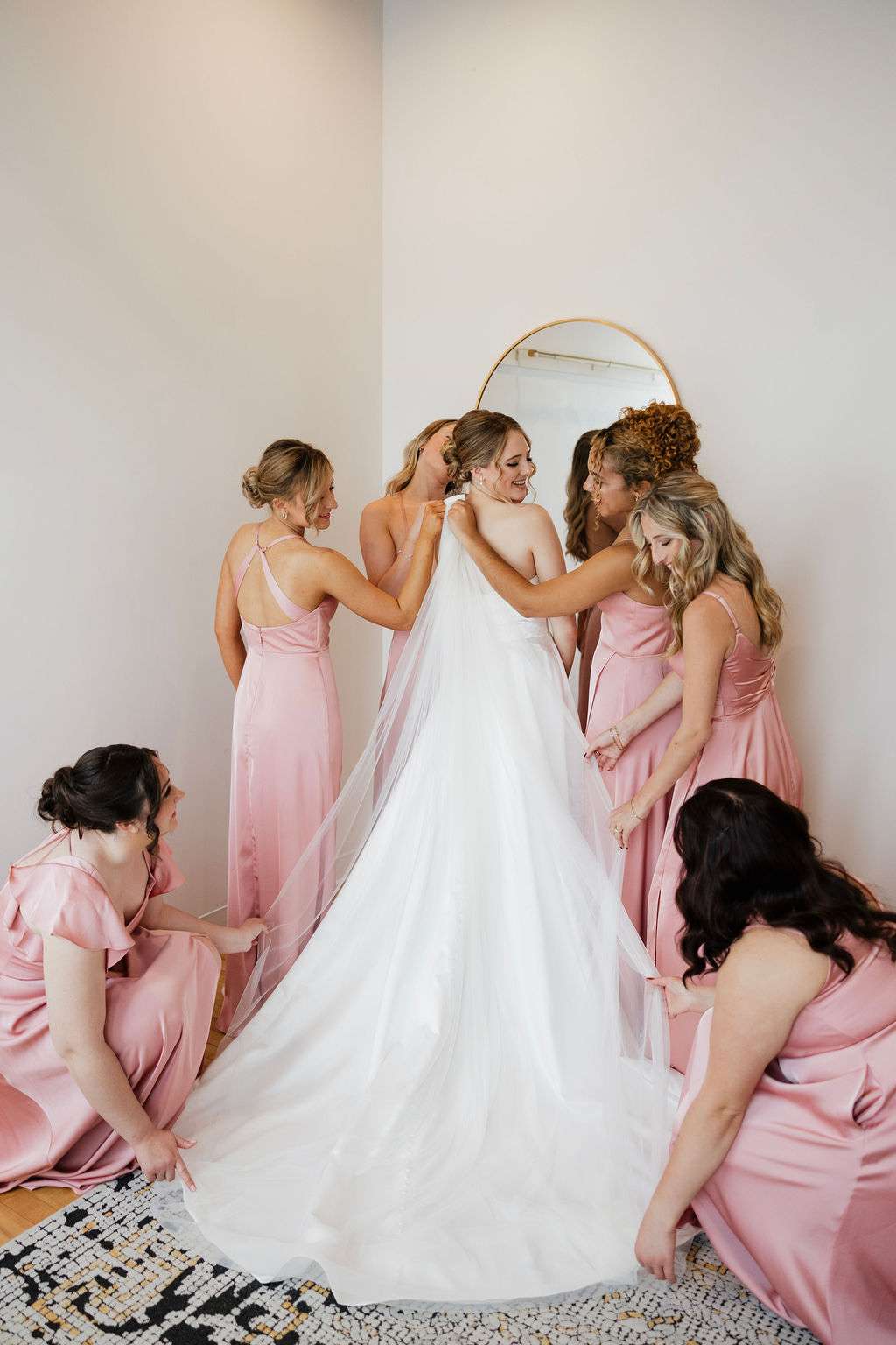 Bride surrounded by bridesmaids in pink dresses helping her with veil in Mercantile Hall’s bridal suite