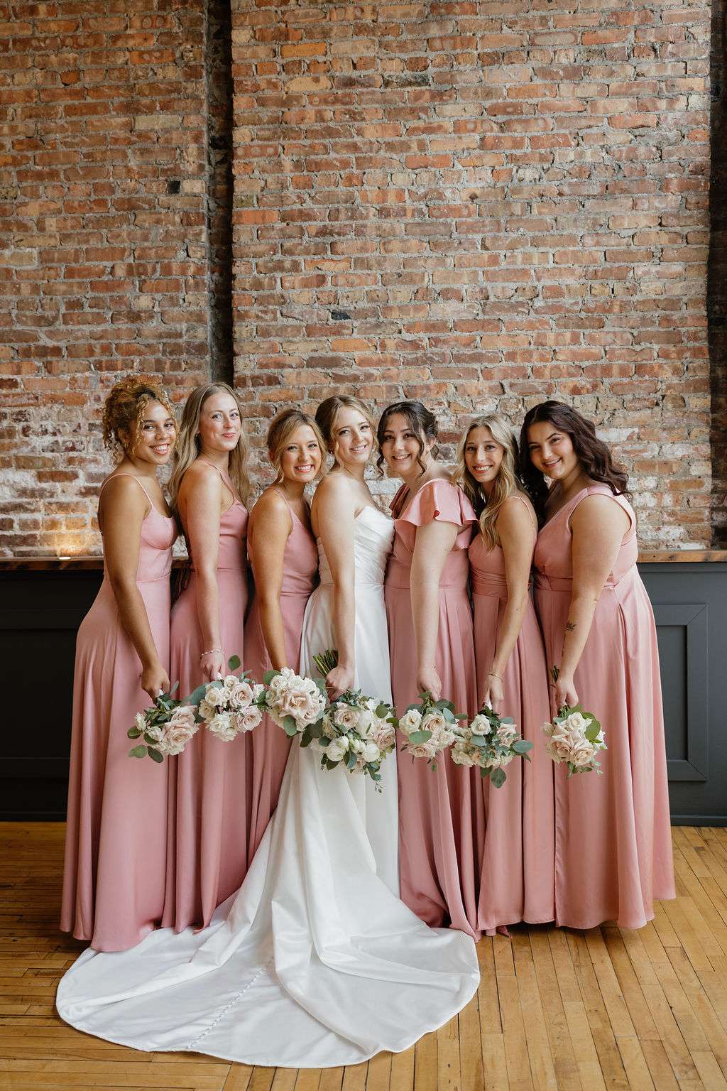 Bride and bridesmaids in blush pink dresses smiling together against brick wall at Mercantile Hall