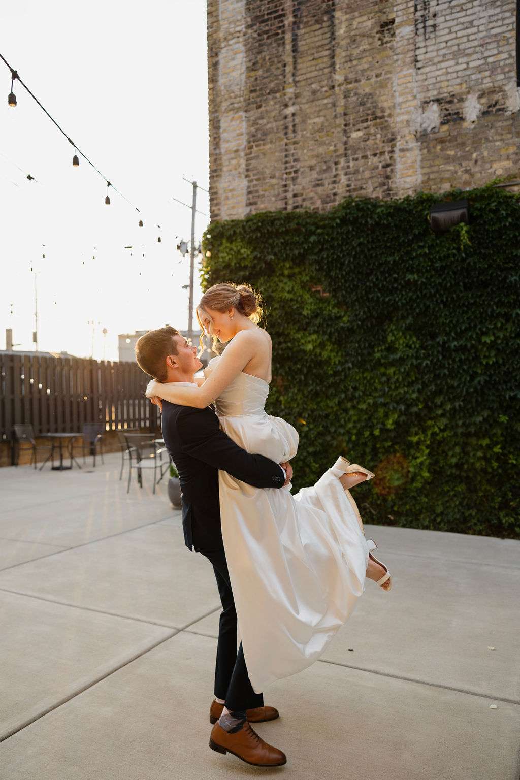 Bride and groom celebrate their wedding at Mercantile Hall in Burlington, WI, with the groom lifting the bride outdoors against a backdrop of ivy-covered brick and string lights.