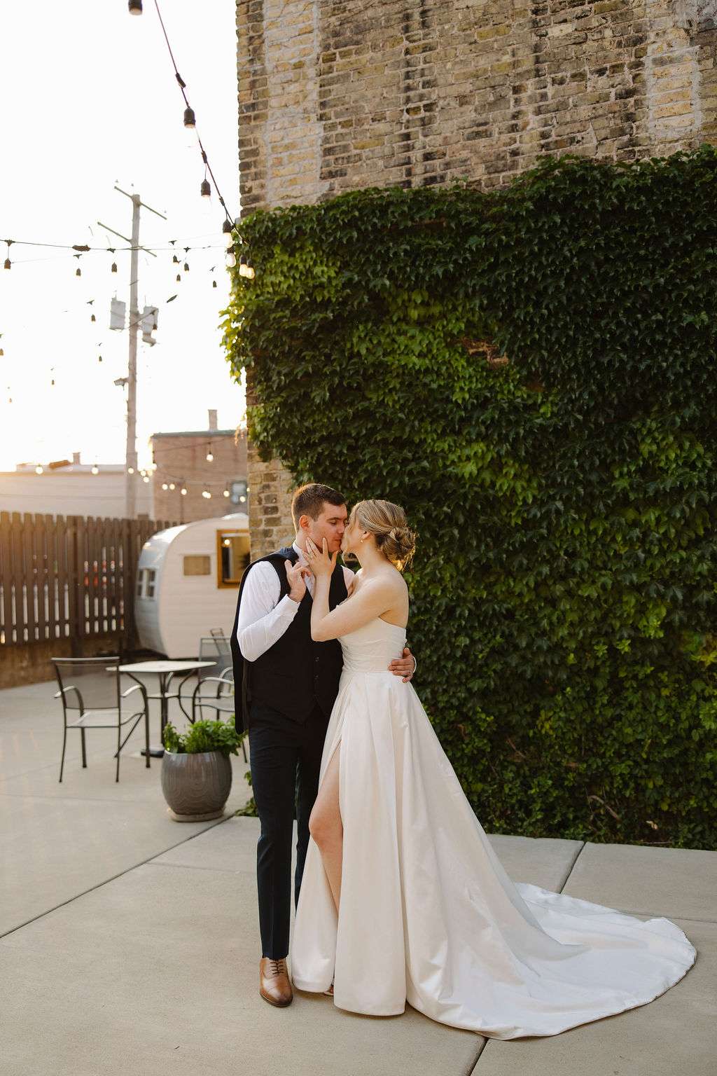 Bride and groom sharing a quiet moment during golden hour in a charming ivy-covered courtyard with string lights and a vintage camper in the background.