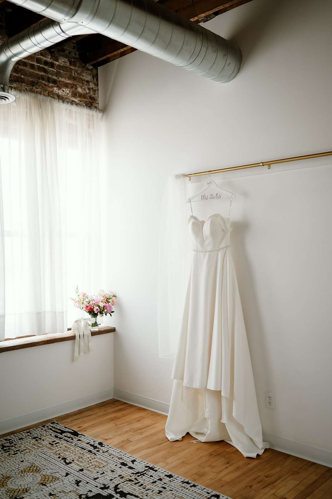 Simple ivory wedding gown hanging from a personalized hanger in a softly lit bridal suite with wood floors and industrial details.