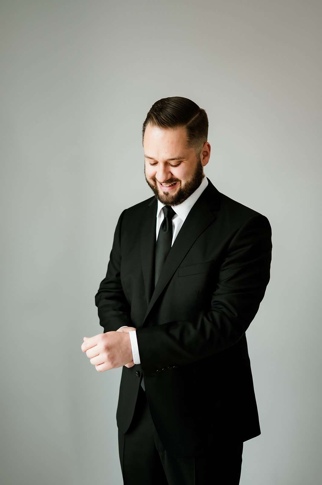 Groom smiling as he adjusts his cufflinks in a sleek black suit against a soft gray background.